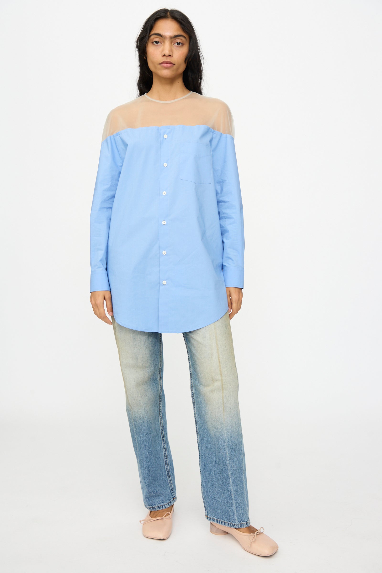 A woman stands facing forward in the MM6 Long Sleeved Top in Sky Blue with a sheer beige yoke, faded blue jeans, and light pink ballet flats against a plain white background. 