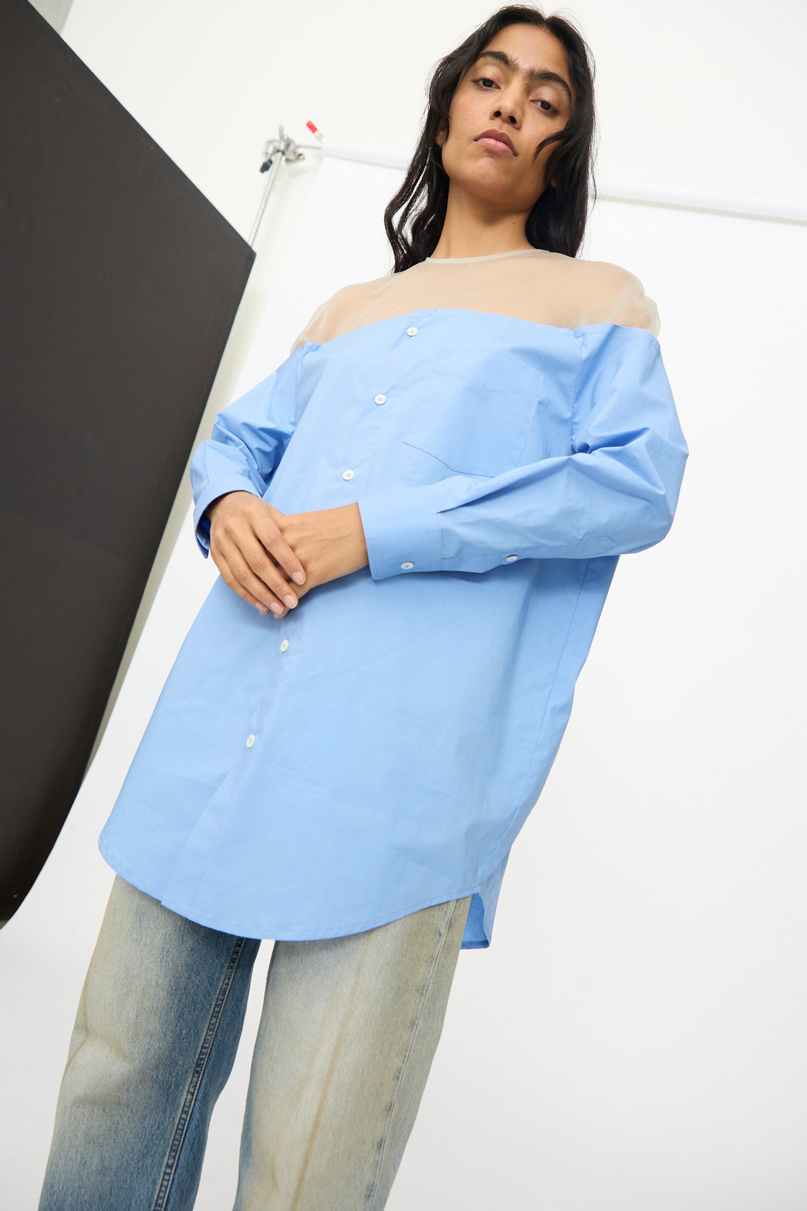 A person stands against a plain studio backdrop wearing the MM6 Long Sleeved Top in Sky Blue with a sheer beige yoke and light-wash jeans.