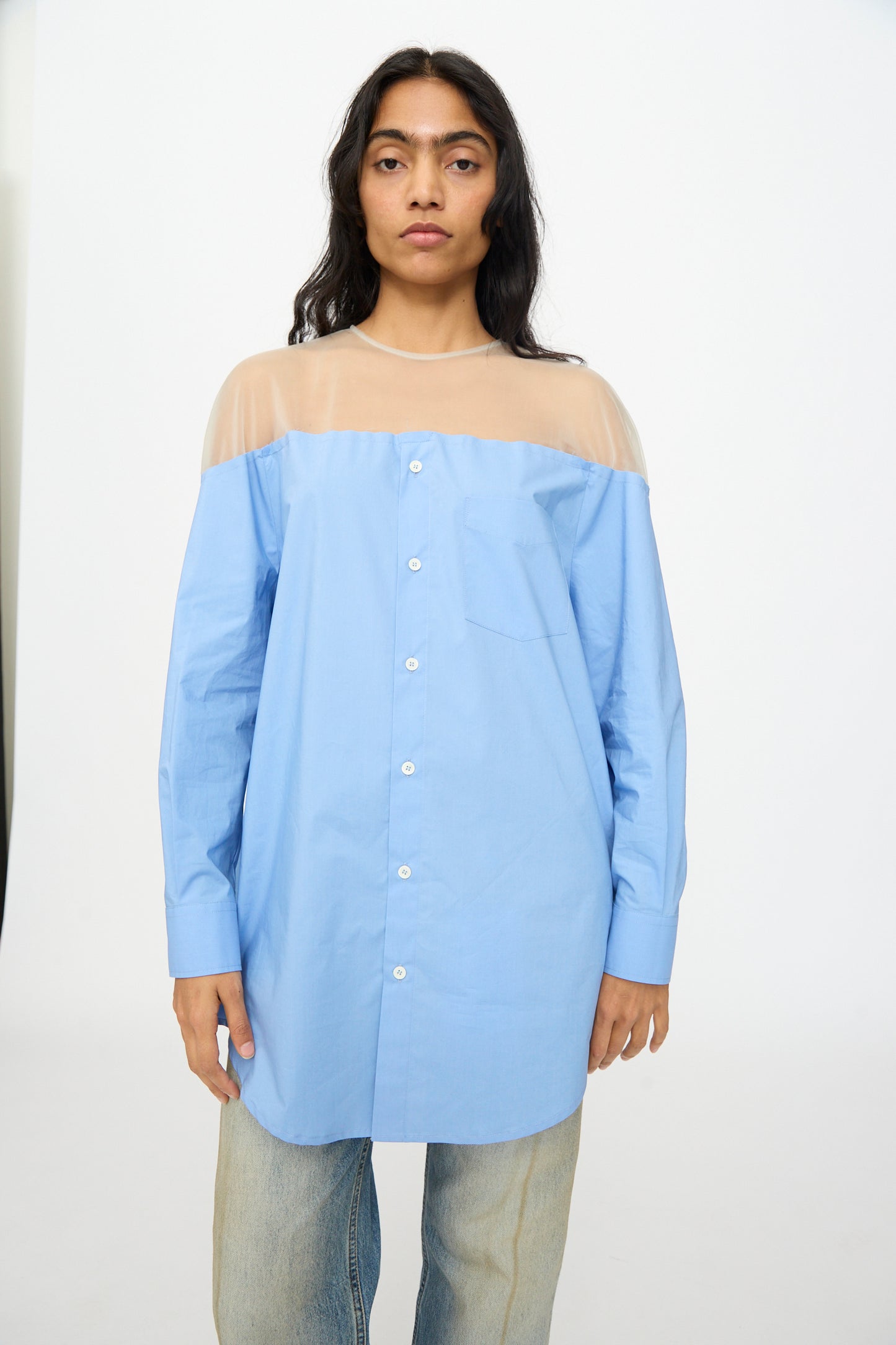 A person wears the MM6 Long Sleeved Top in Sky Blue, featuring sheer beige shoulder panels, paired with light blue jeans, and stands against a plain white background.