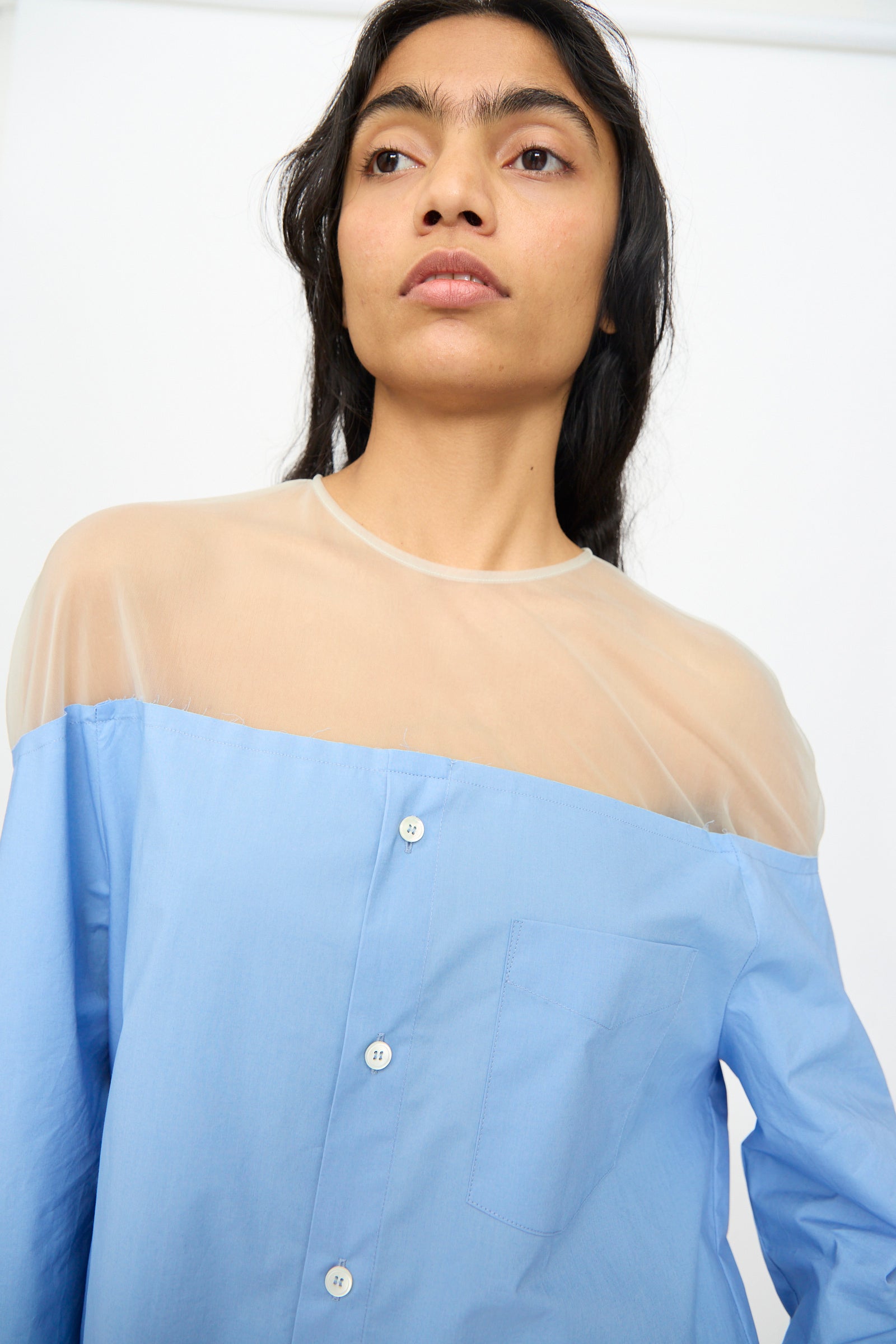 A model wears the MM6 Long Sleeved Top in Sky Blue, featuring a sheer beige mesh panel, and gazes slightly upward against a plain background.