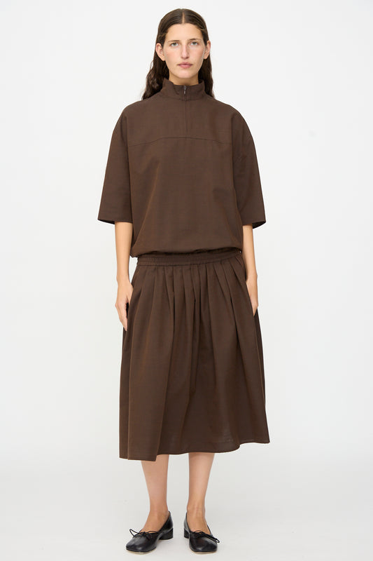 A person stands facing forward, wearing the MM6 Maxi Dress in Brown—a loose-fitting piece featuring a high collar and elbow-length sleeves, crafted from a luxurious wool-cotton blend. They have long hair and wear black shoes.
