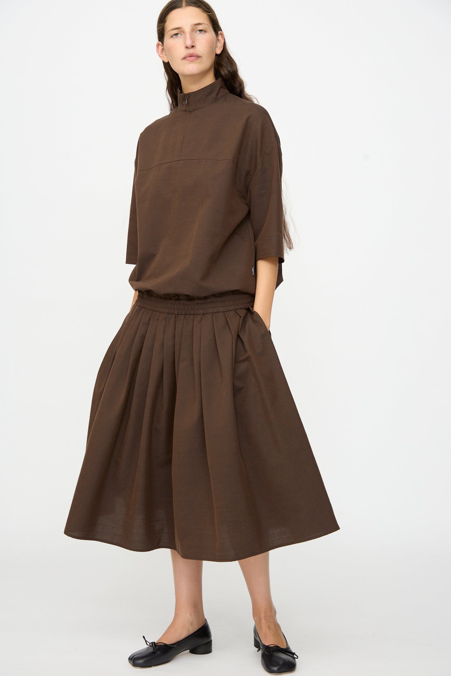 A woman stands against a plain background wearing the MM6 Maxi Dress in Brown, crafted in Italy from a luxurious wool-cotton blend. This elegant dress features three-quarter sleeves and pleated details, exemplifying the brand’s commitment to timeless style and quality craftsmanship. She pairs the look with classic black ballet flats.