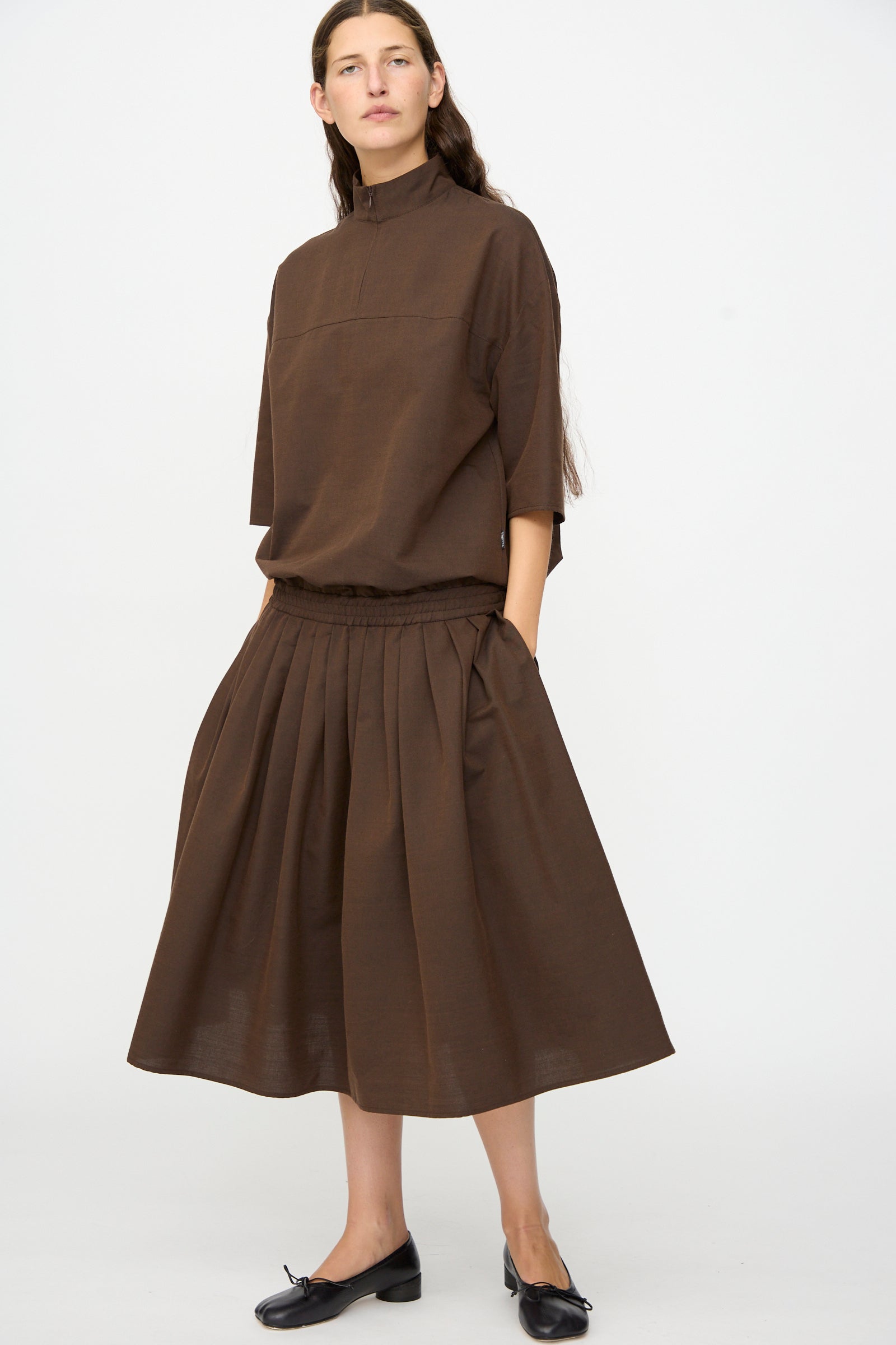 A woman stands against a plain background wearing the MM6 Maxi Dress in Brown, crafted in Italy from a luxurious wool-cotton blend. This elegant dress features three-quarter sleeves and pleated details, exemplifying the brand’s commitment to timeless style and quality craftsmanship. She pairs the look with classic black ballet flats.