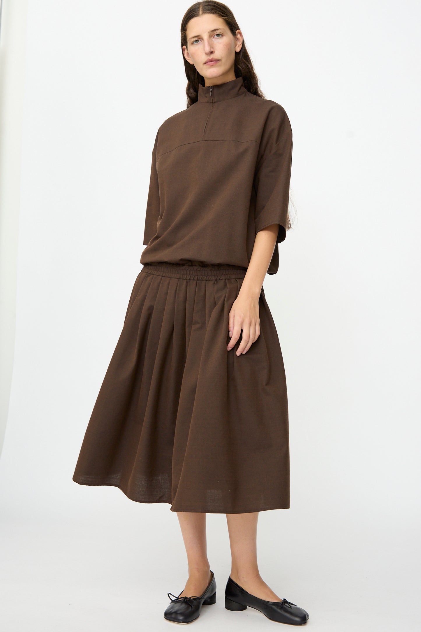 A person stands against a plain white background wearing the MM6 Maxi Dress in Brown, a loose, knee-length dress with a high collar crafted from a luxurious wool-cotton blend, paired with black flat shoes.
