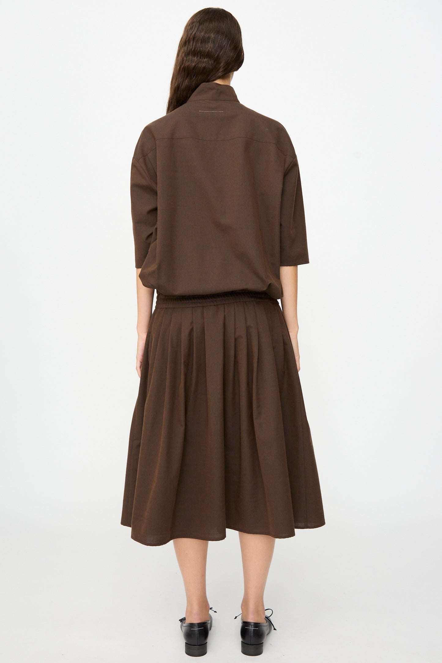 A person stands with their back to the camera, wearing the MM6 Maxi Dress in Brown, a loose-fitting piece crafted from a soft wool-cotton blend with elbow-length sleeves and subtle pleating. Their long, wavy hair flows down their back.