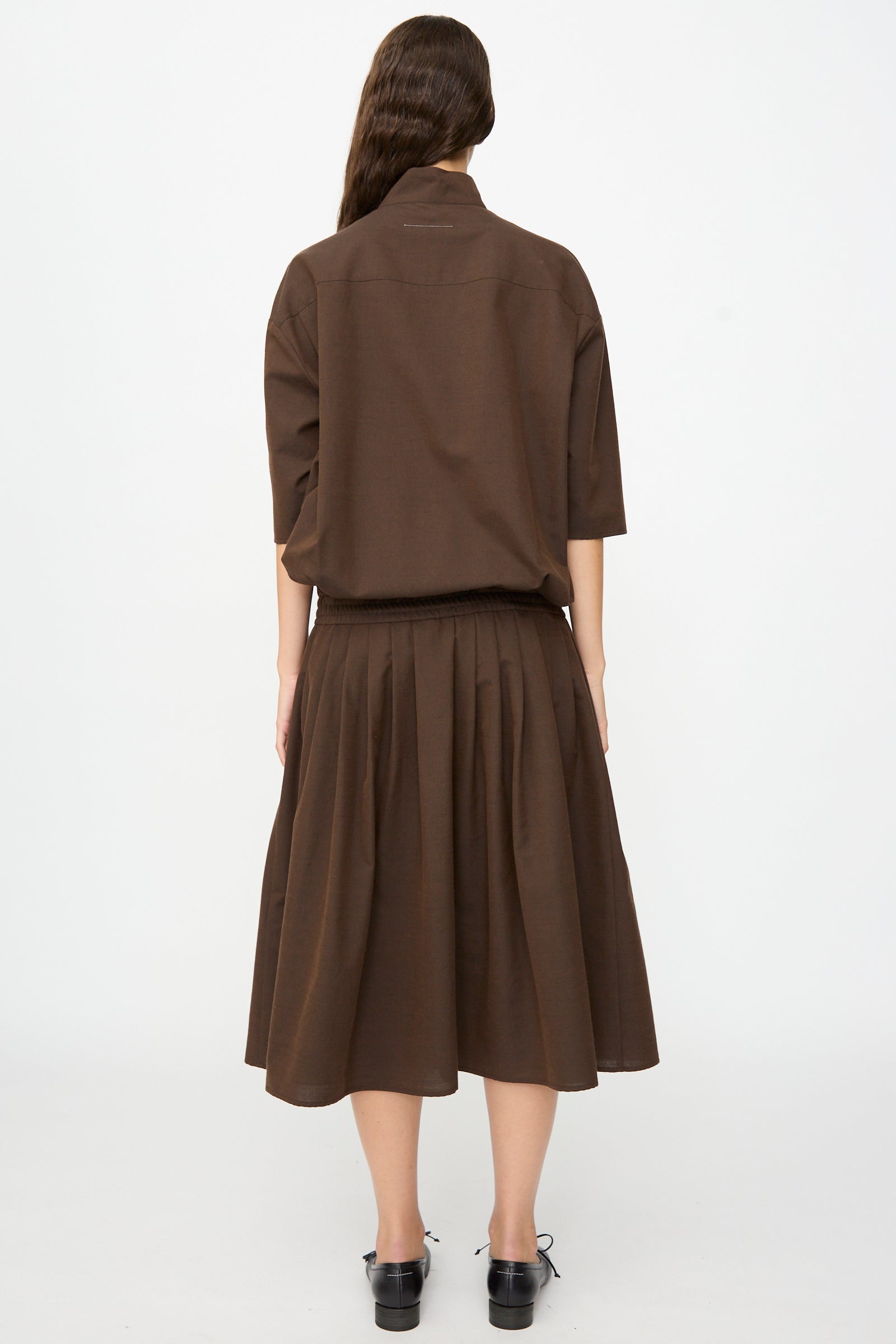A person stands with their back to the camera, wearing the MM6 Maxi Dress in Brown, a loose-fitting piece crafted from a soft wool-cotton blend with elbow-length sleeves and subtle pleating. Their long, wavy hair flows down their back.