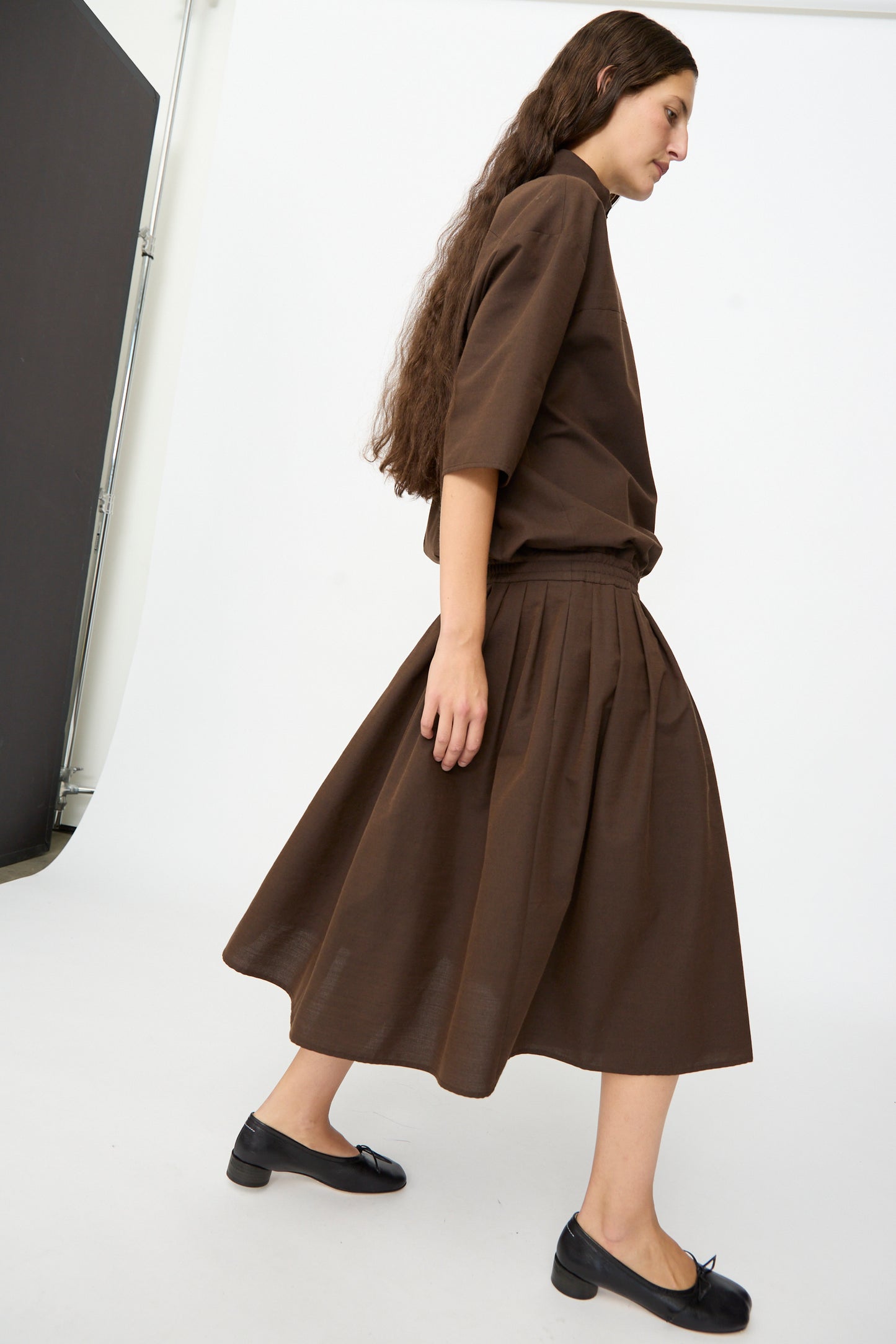 A person with long brown hair is wearing the MM6 Maxi Dress in Brown, paired with black flat shoes and walking against a plain indoor background.