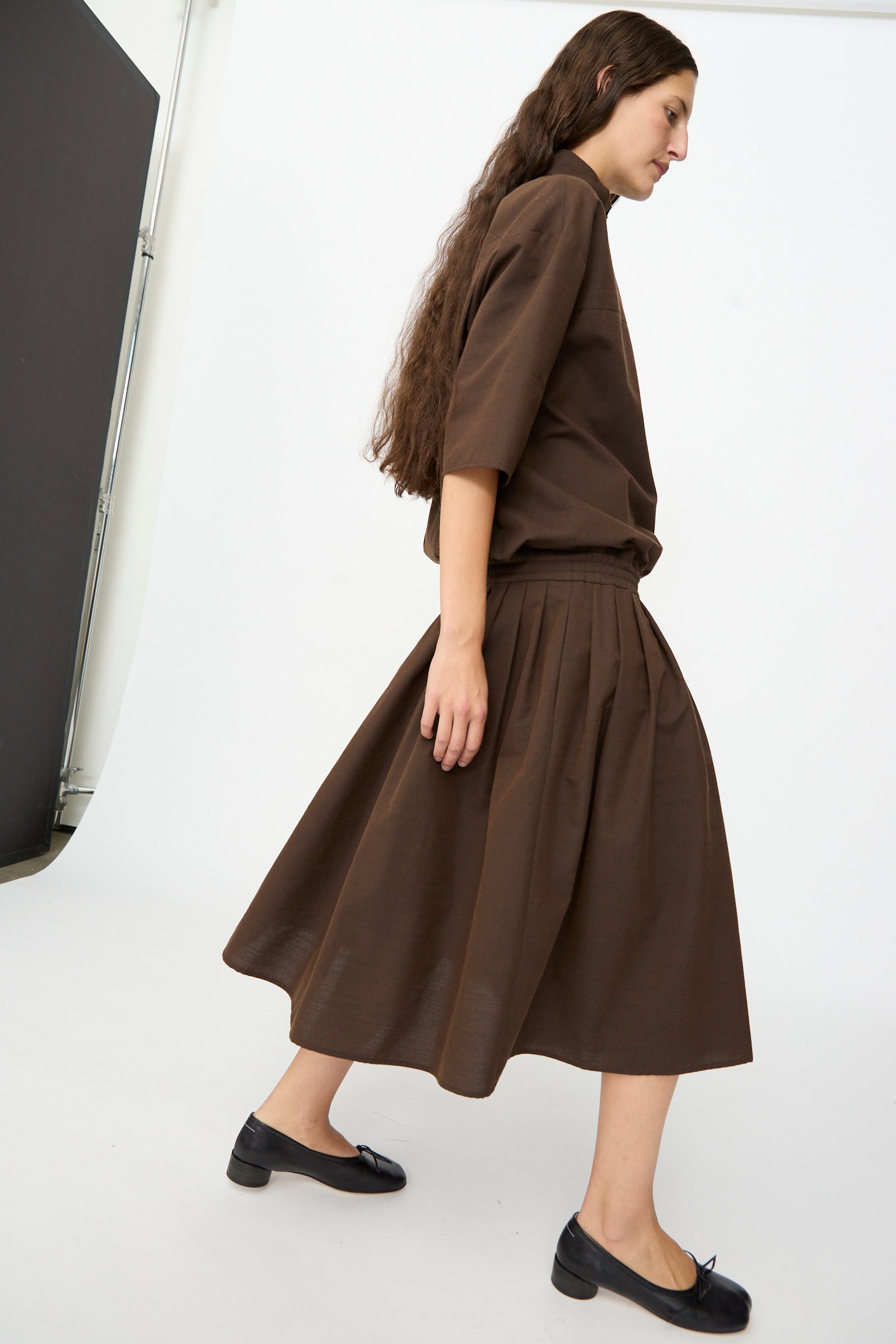 A person with long brown hair is wearing the MM6 Maxi Dress in Brown, paired with black flat shoes and walking against a plain indoor background.