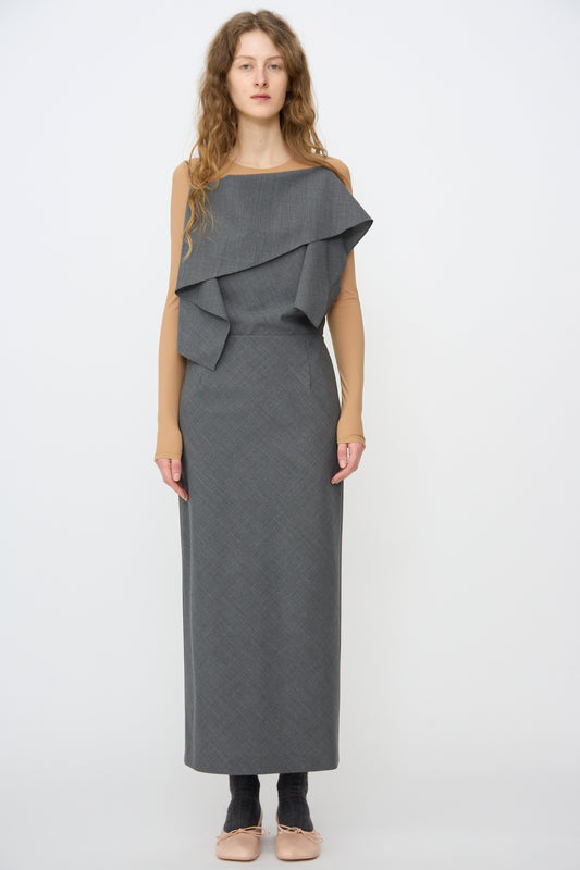 A person stands facing forward in the MM6 Maxi Dress in Dark Grey Melange, featuring an asymmetrical neckline, layered detailing, and nude long sleeves, set against a plain white background.