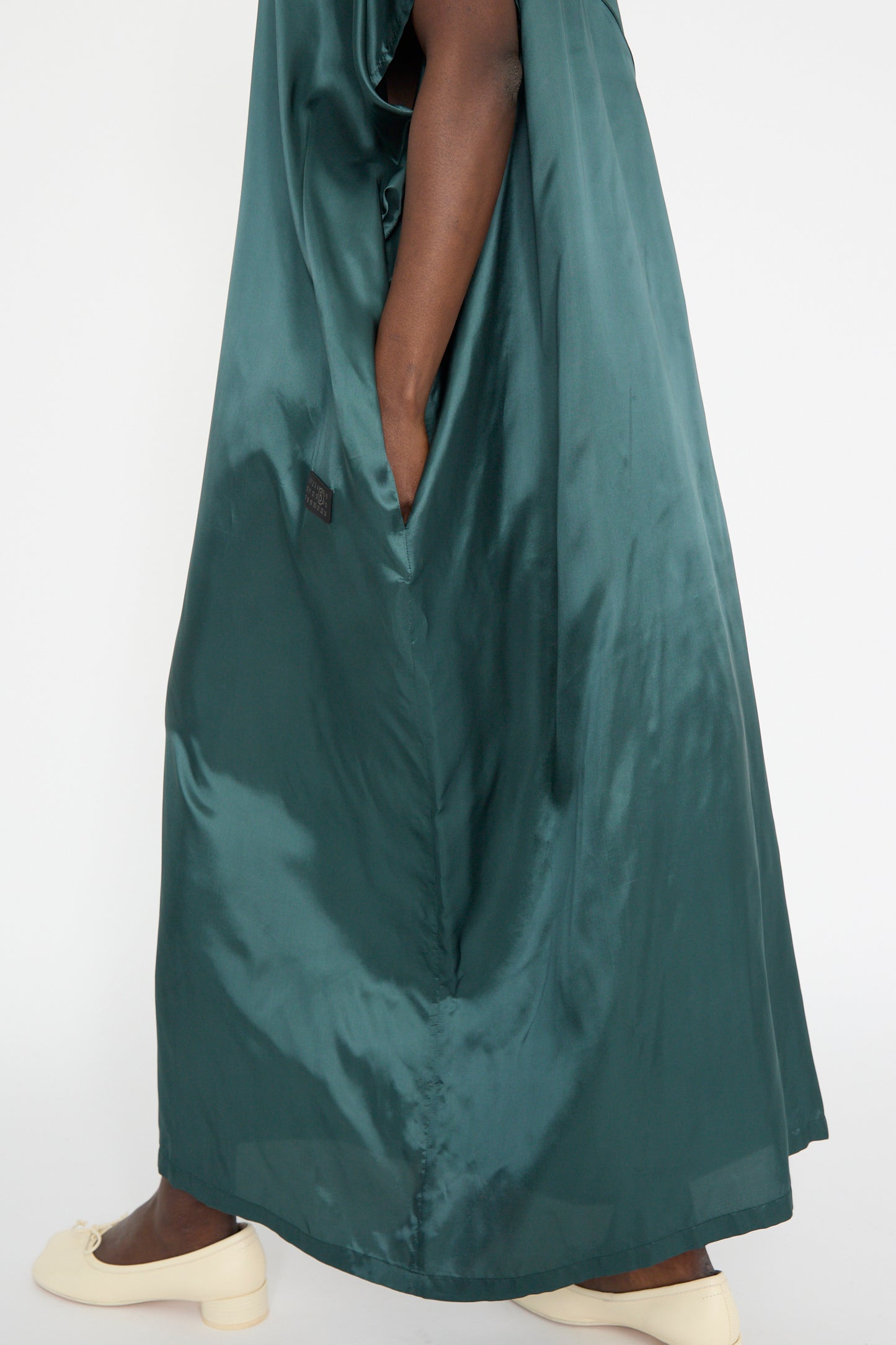 A person in an oversized fit MM6 Maxi Dress in green satin twill, hands in pockets, paired with cream flat shoes.