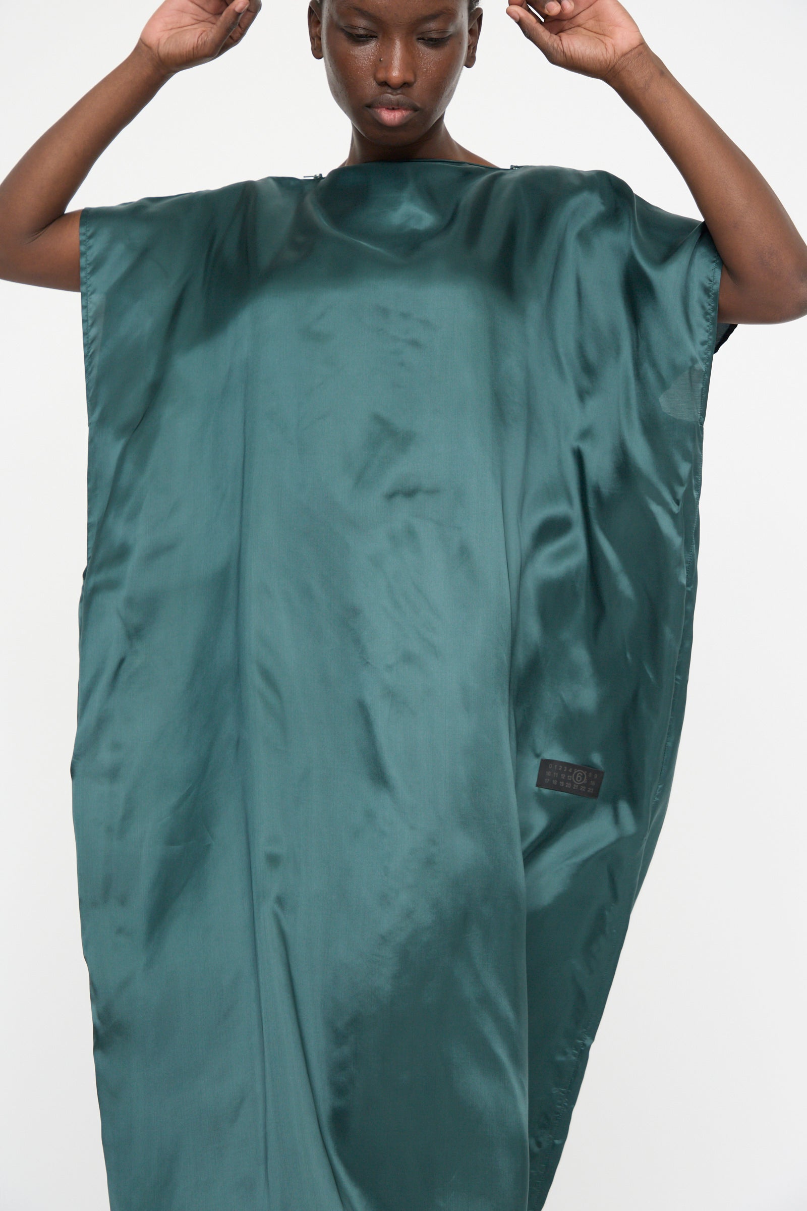 A person is wearing the MM6 Maxi Dress in Green, a loose-fitting, oversized satin twill garment featuring a side label.