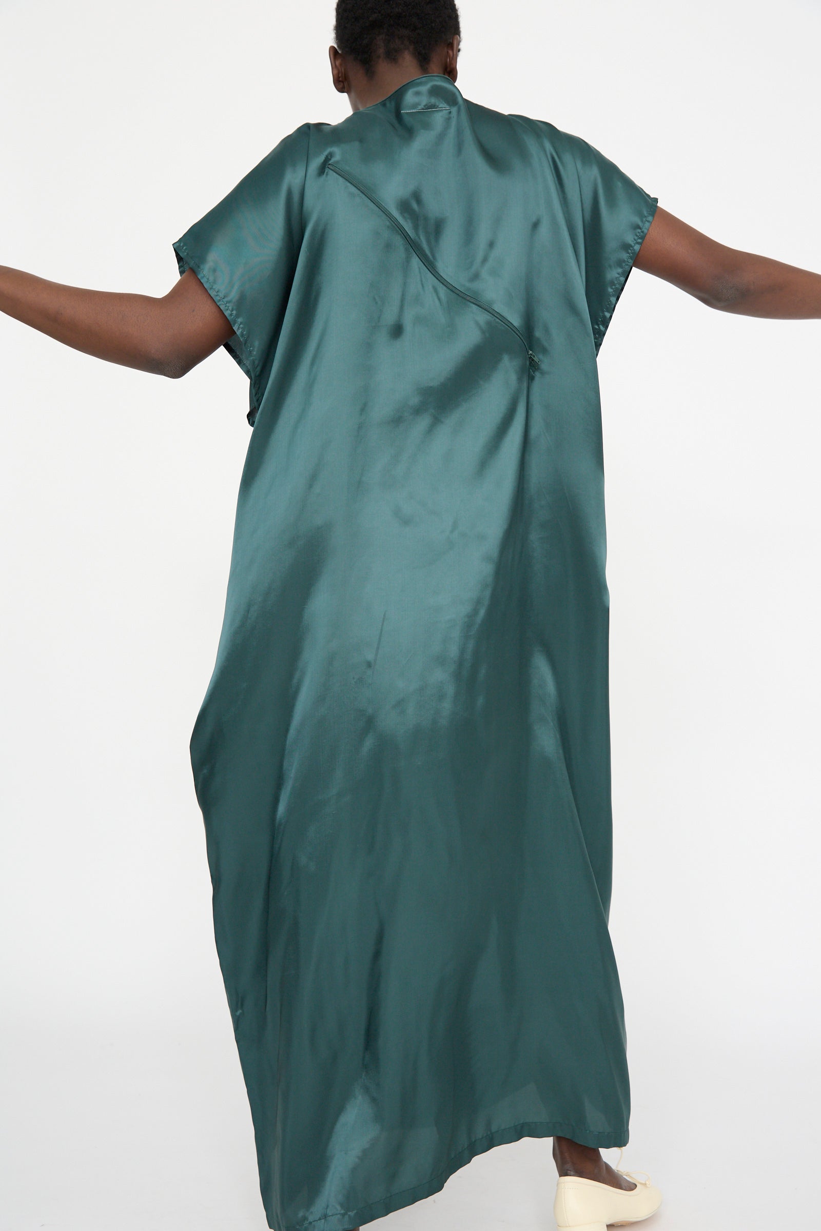 A person stands with arms slightly extended, wearing the Maxi Dress in Green by MM6. They face away from the camera against a plain white background.