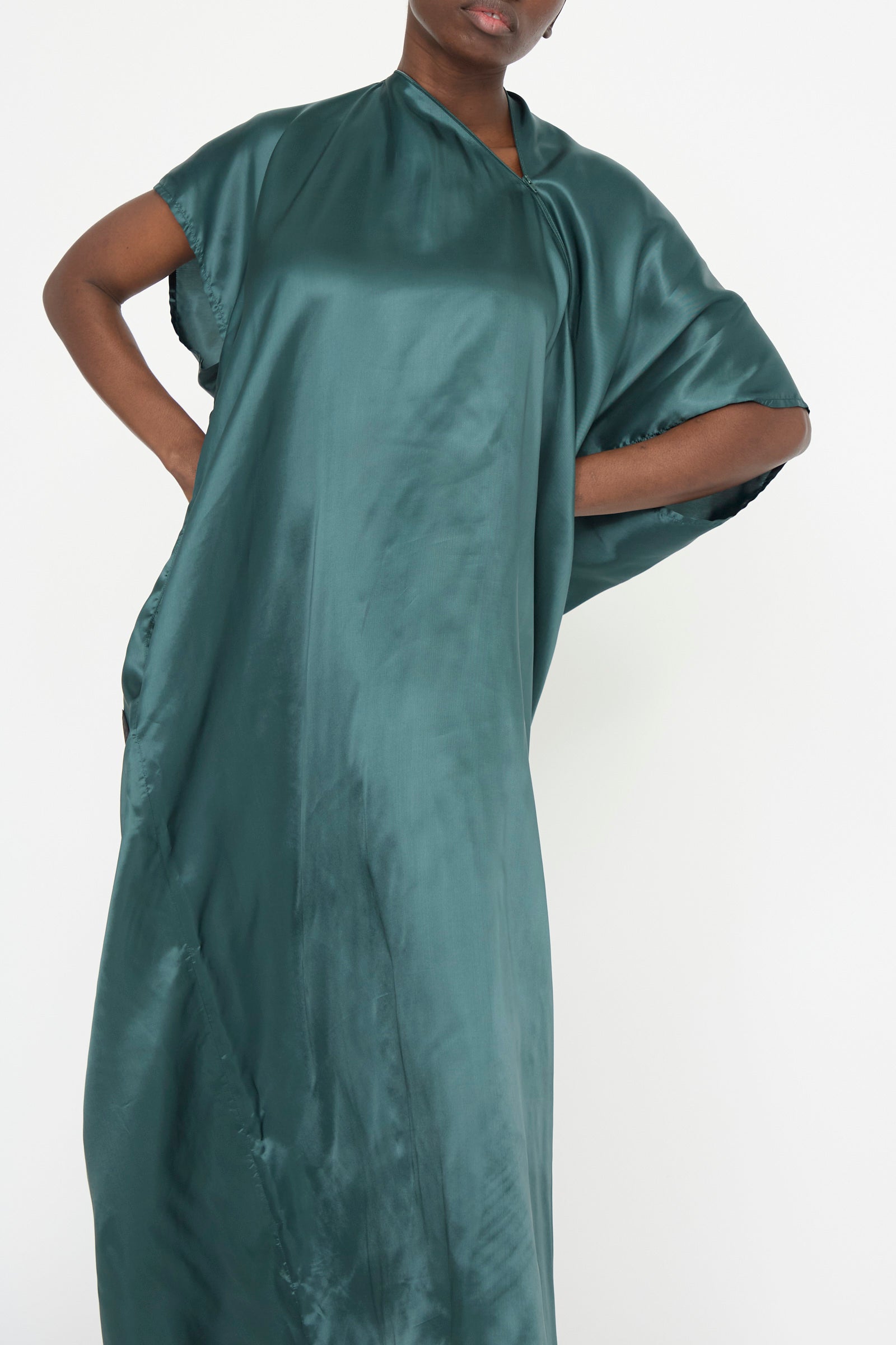 A person models MM6's Maxi Dress in Green, featuring a shiny satin twill fabric with a flowing design, standing with one hand on their hip against a plain white background.