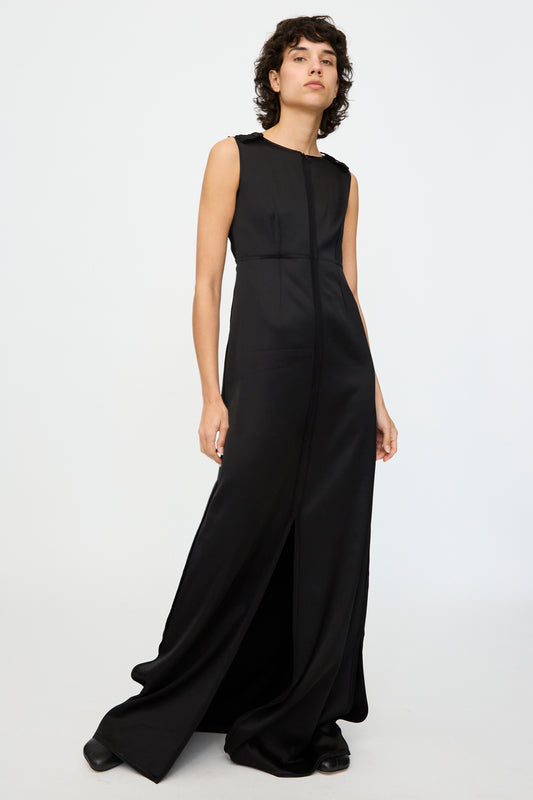 A person wears an MM6 sleeveless black Maxi Dress featuring shoulder buttons and a front seam, paired with black shoes, against a white background.