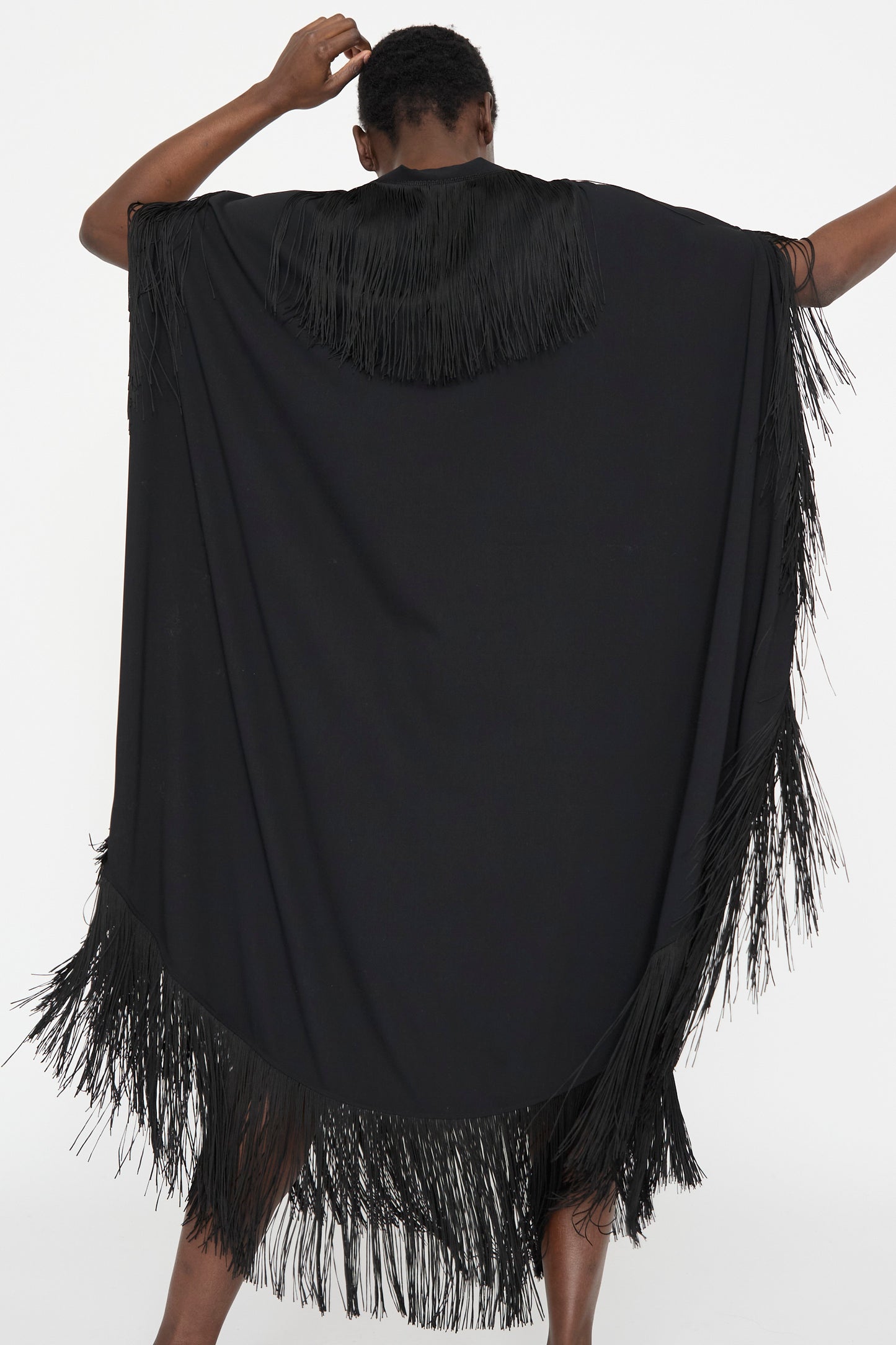 A person wearing MM6's Midi Dress in black, featuring fringe details, stands with their back to the camera against a plain backdrop.