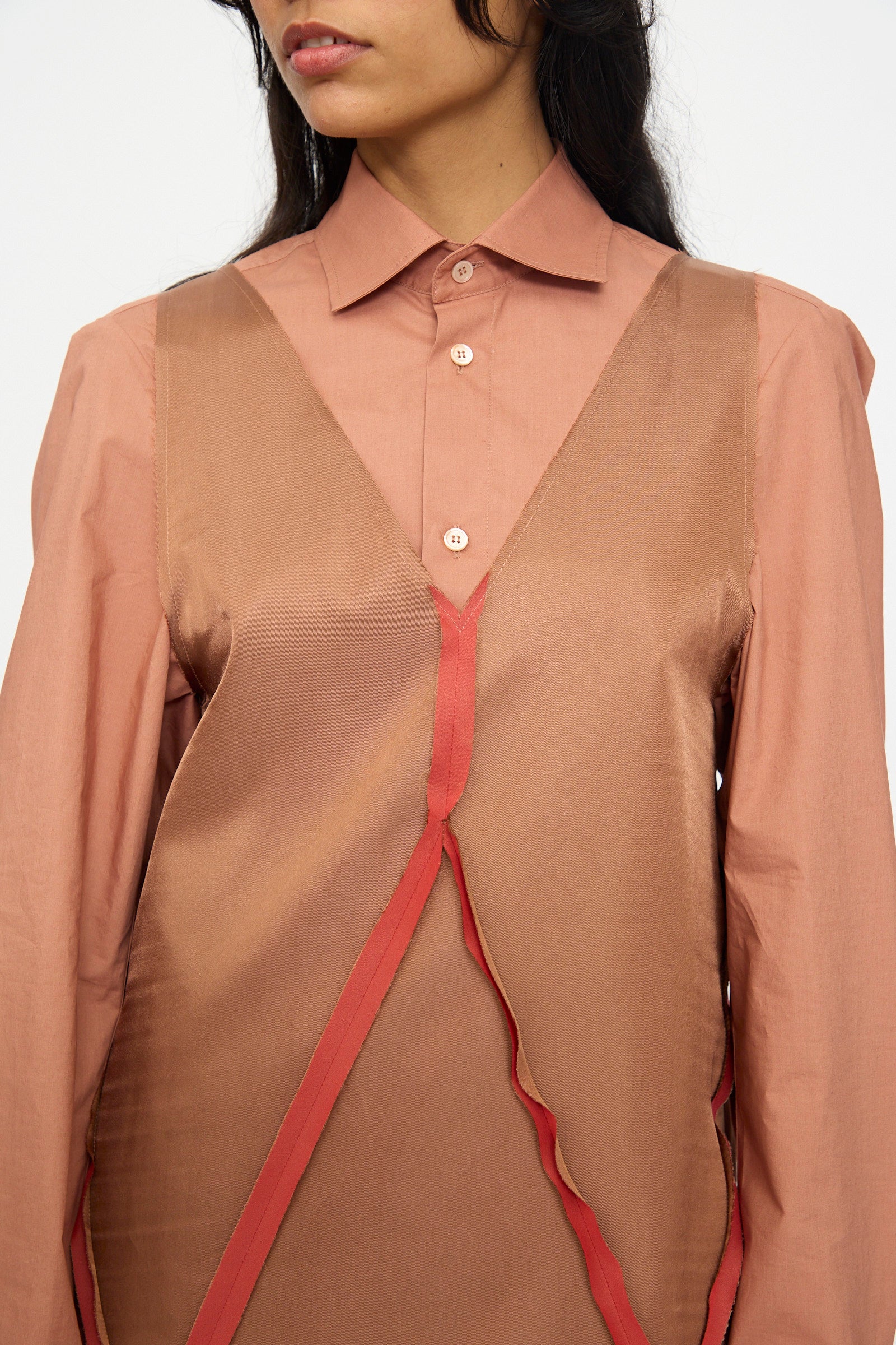 A woman wears the MM6 Midi Dress in Light Brown—a sleeveless, V-neck style with asymmetrical red trim—layered over a buttoned, collared, long-sleeve shirt. Only her upper body and face are partially visible.