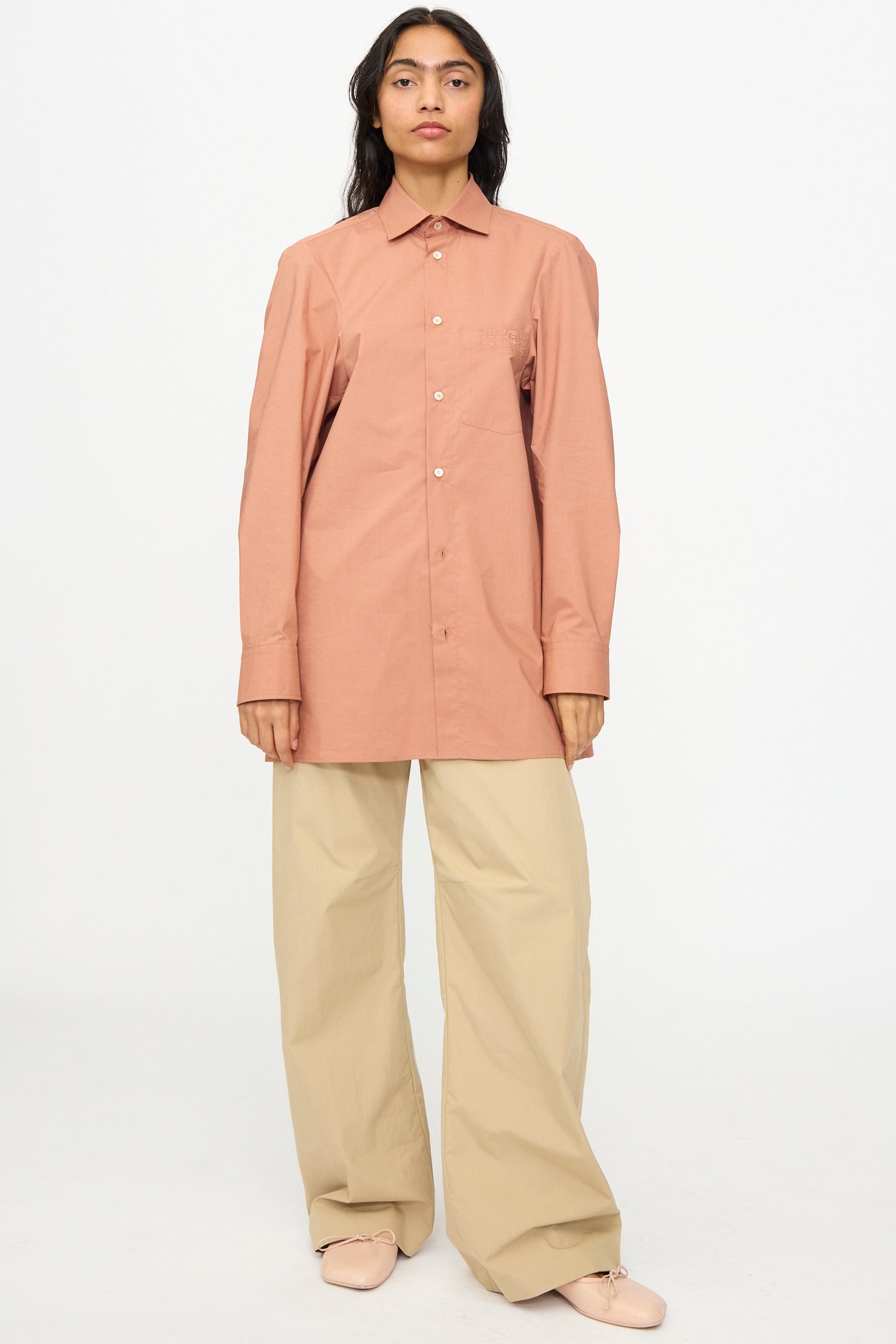 A person stands against a white background, wearing MM6's Pants in Beige paired with a long-sleeve, button-up, salmon-colored shirt.