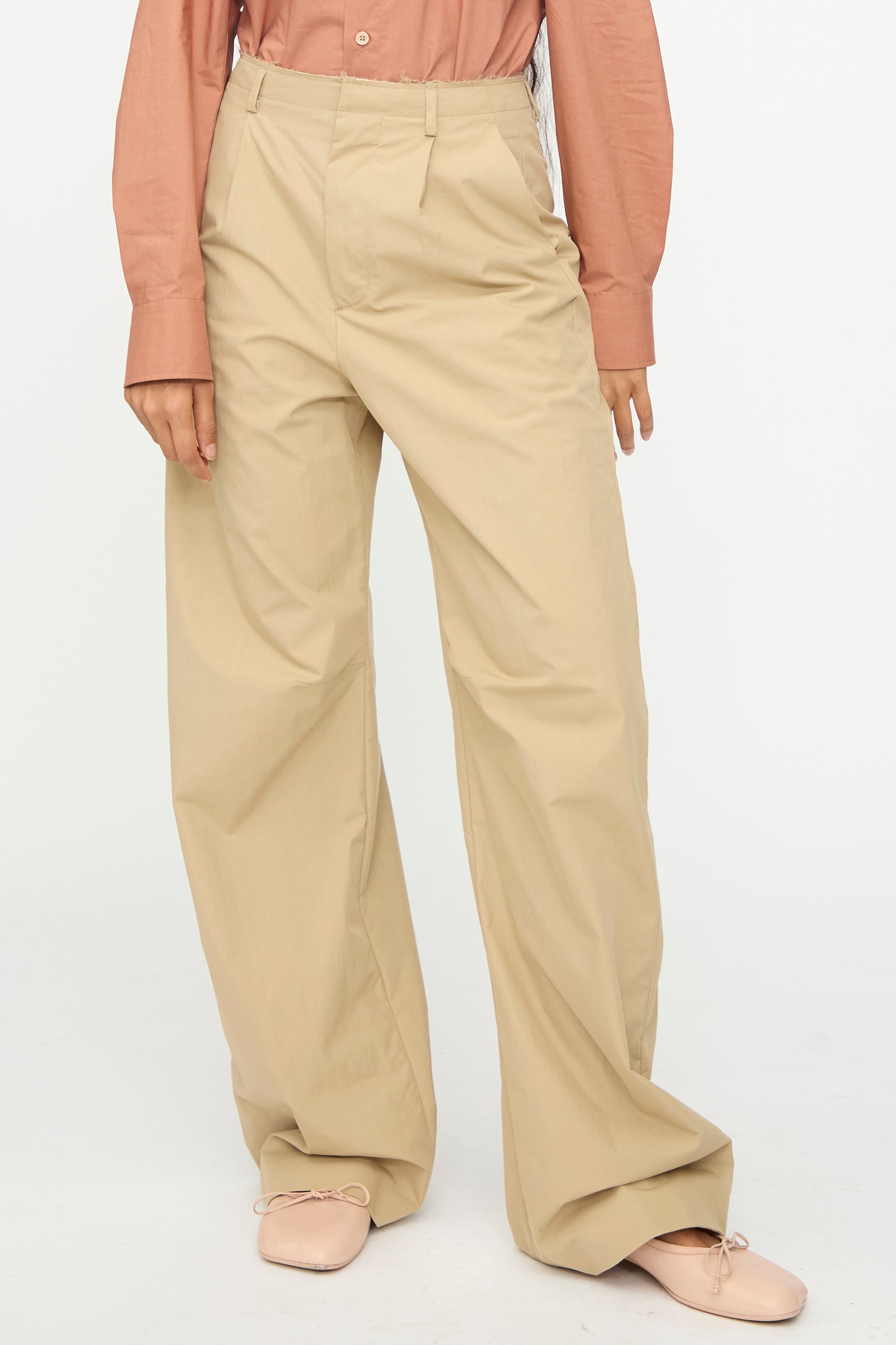 Person wears MM6 Pants in Beige, a muted pink long-sleeve shirt, and pale pink ballet flats, standing against a plain white background.