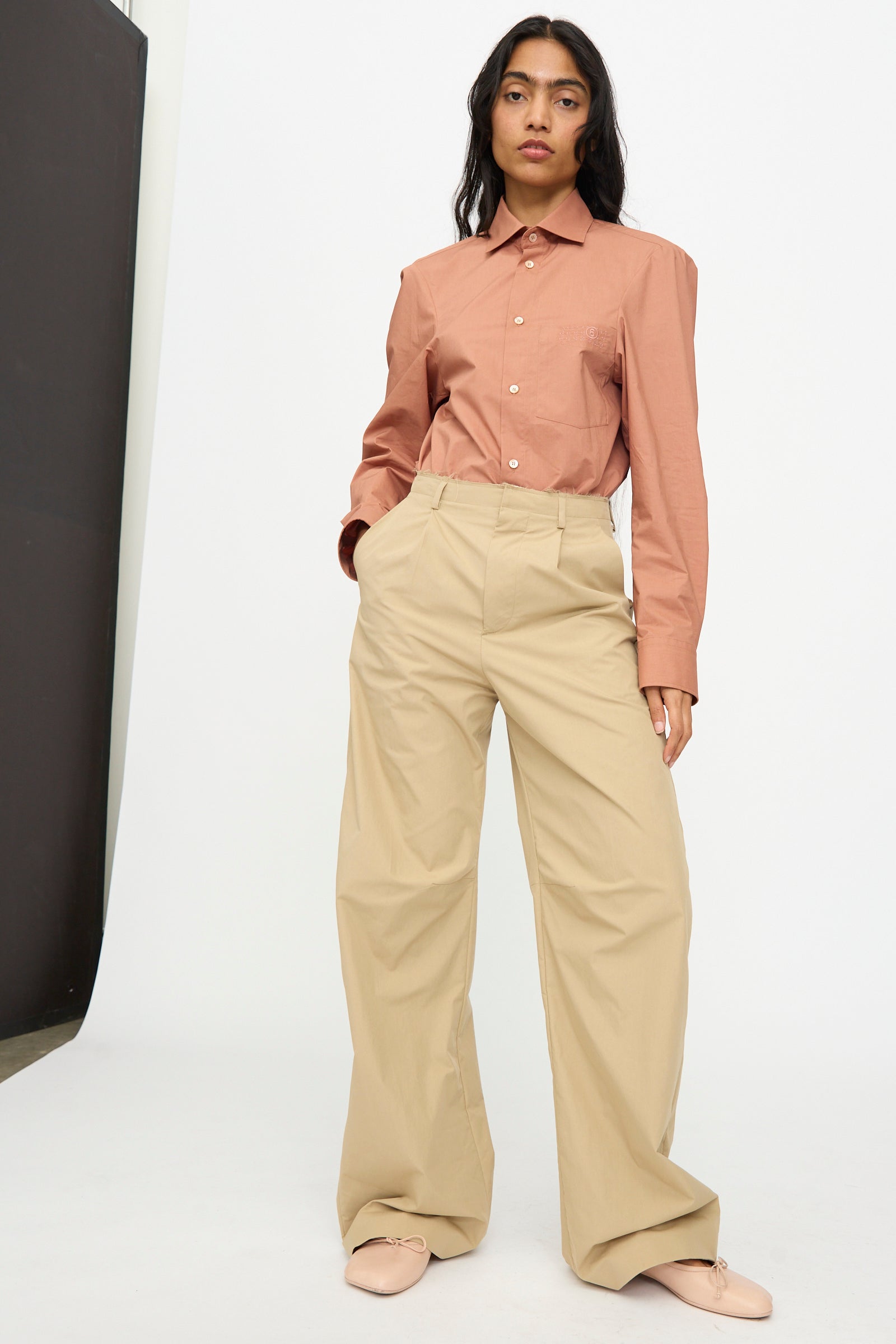 A woman stands against a white background wearing a rust-colored button-up shirt, MM6 Pants in Beige with a wide-leg relaxed fit, and light pink shoes.