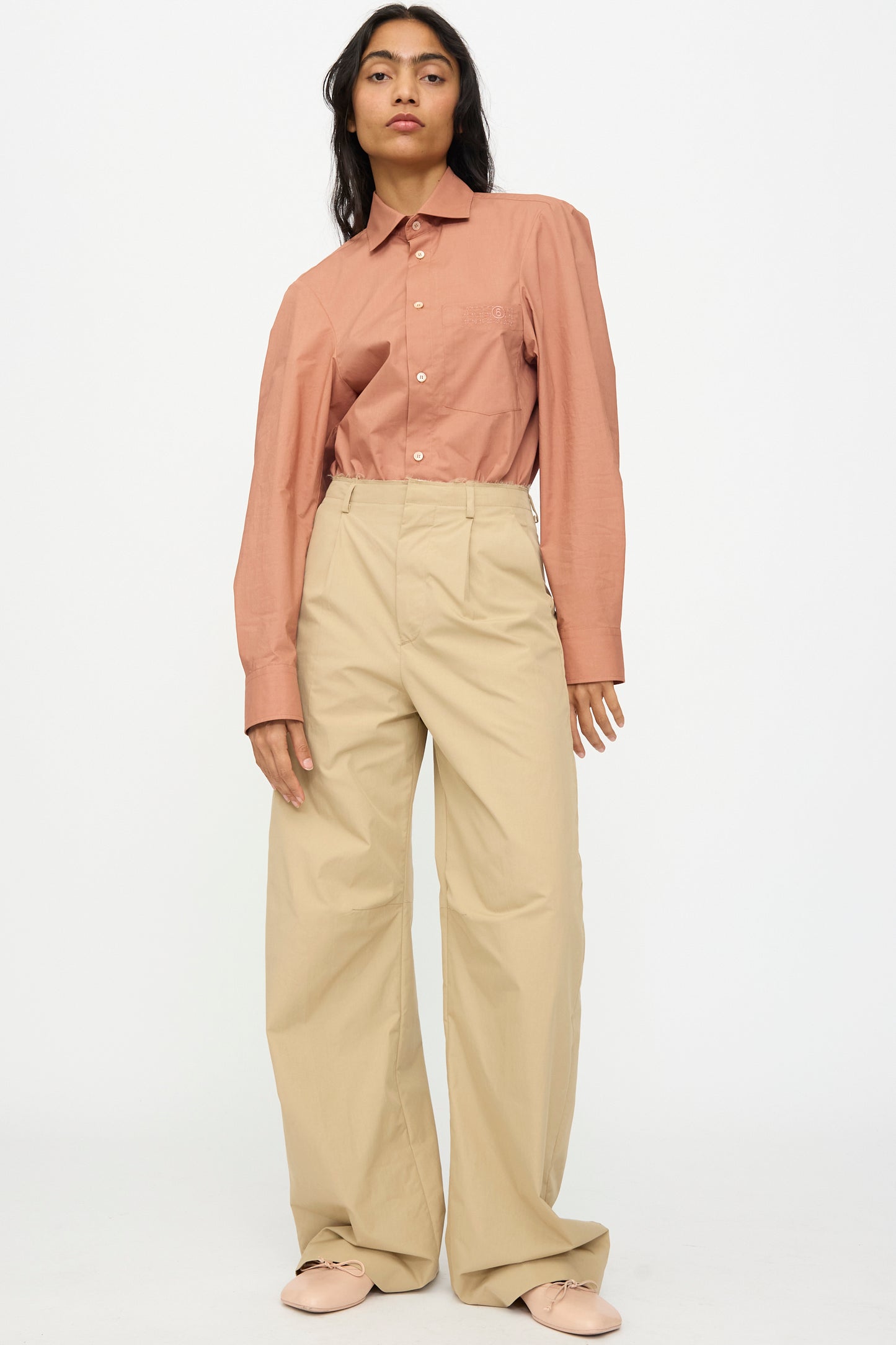 A person stands against a plain background wearing rust-colored button-up shirt, MM6 Pants in Beige (high-waisted relaxed fit wide leg), and light pink shoes.