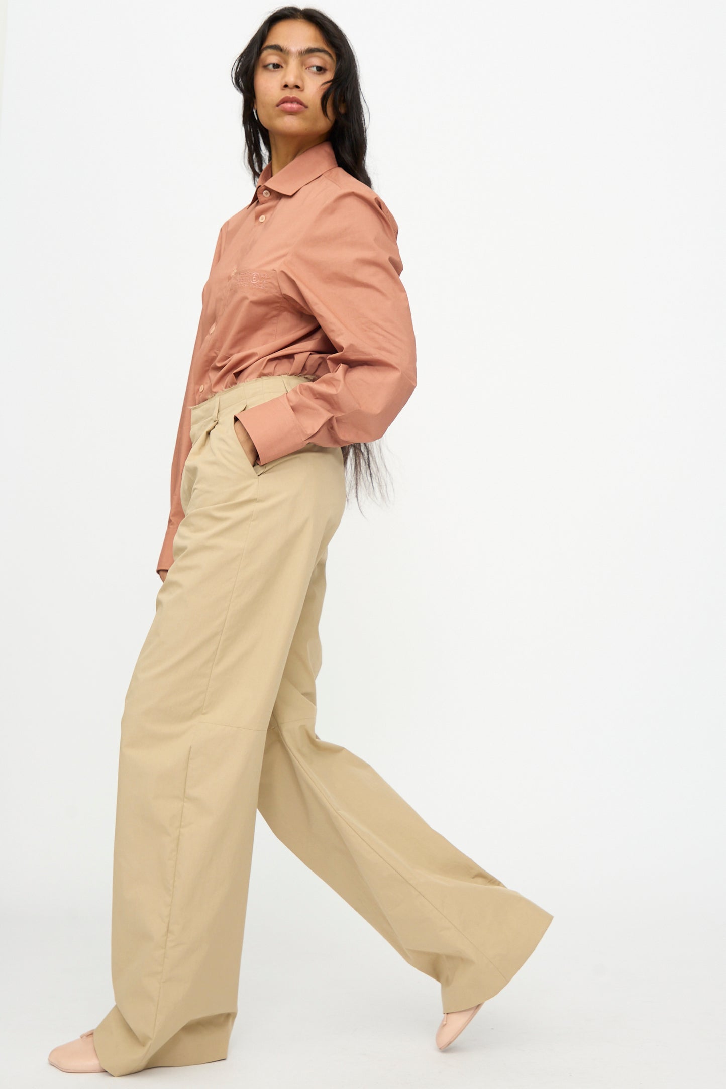 A person wears MM6’s Pants in Beige, styled with a light brown long-sleeve shirt, standing sideways with one hand in their pocket against a plain white background.