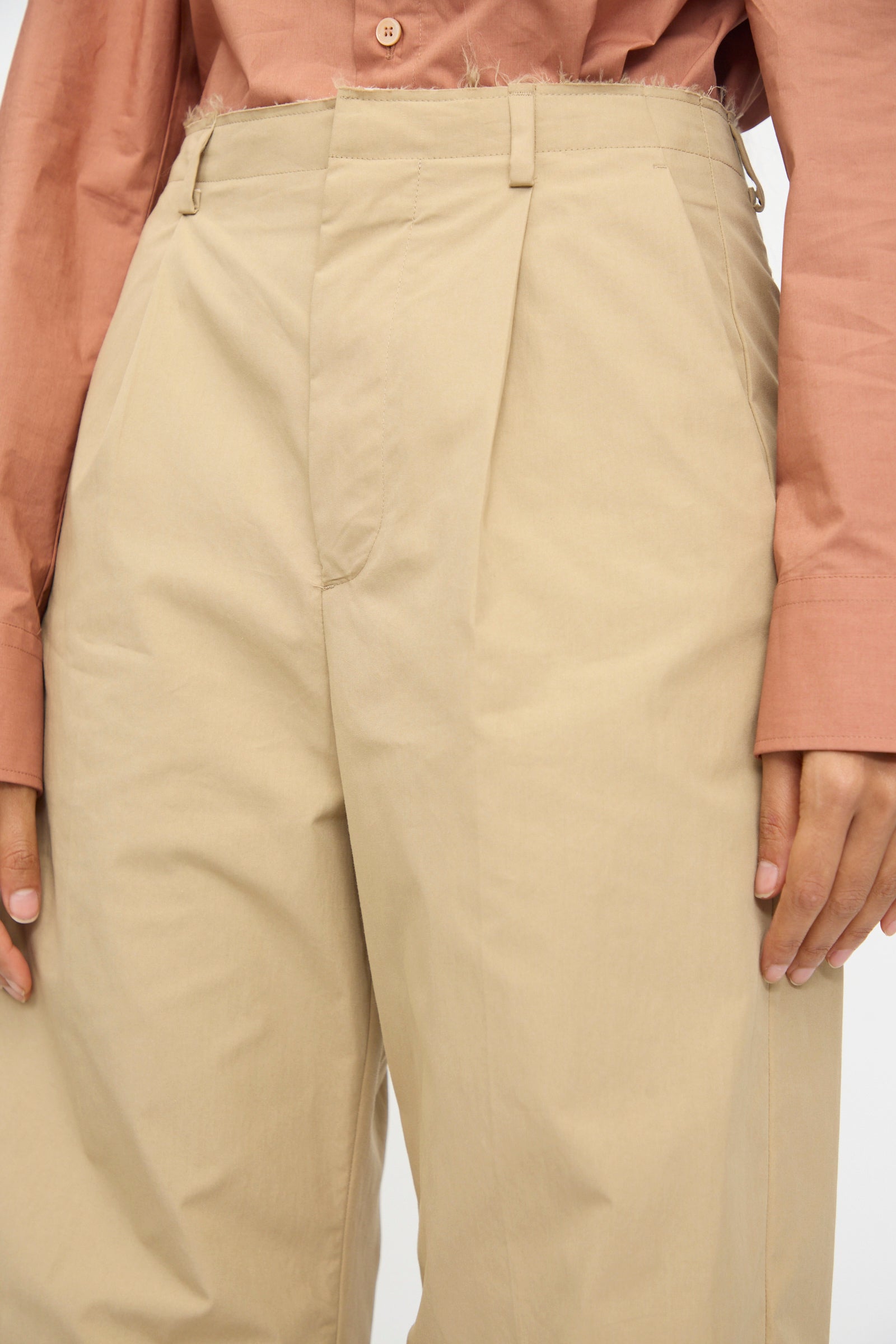 A person wearing a brown button-up shirt with frayed hem and MM6 Pants in Beige—high-waisted, wide-leg, and pleated.
