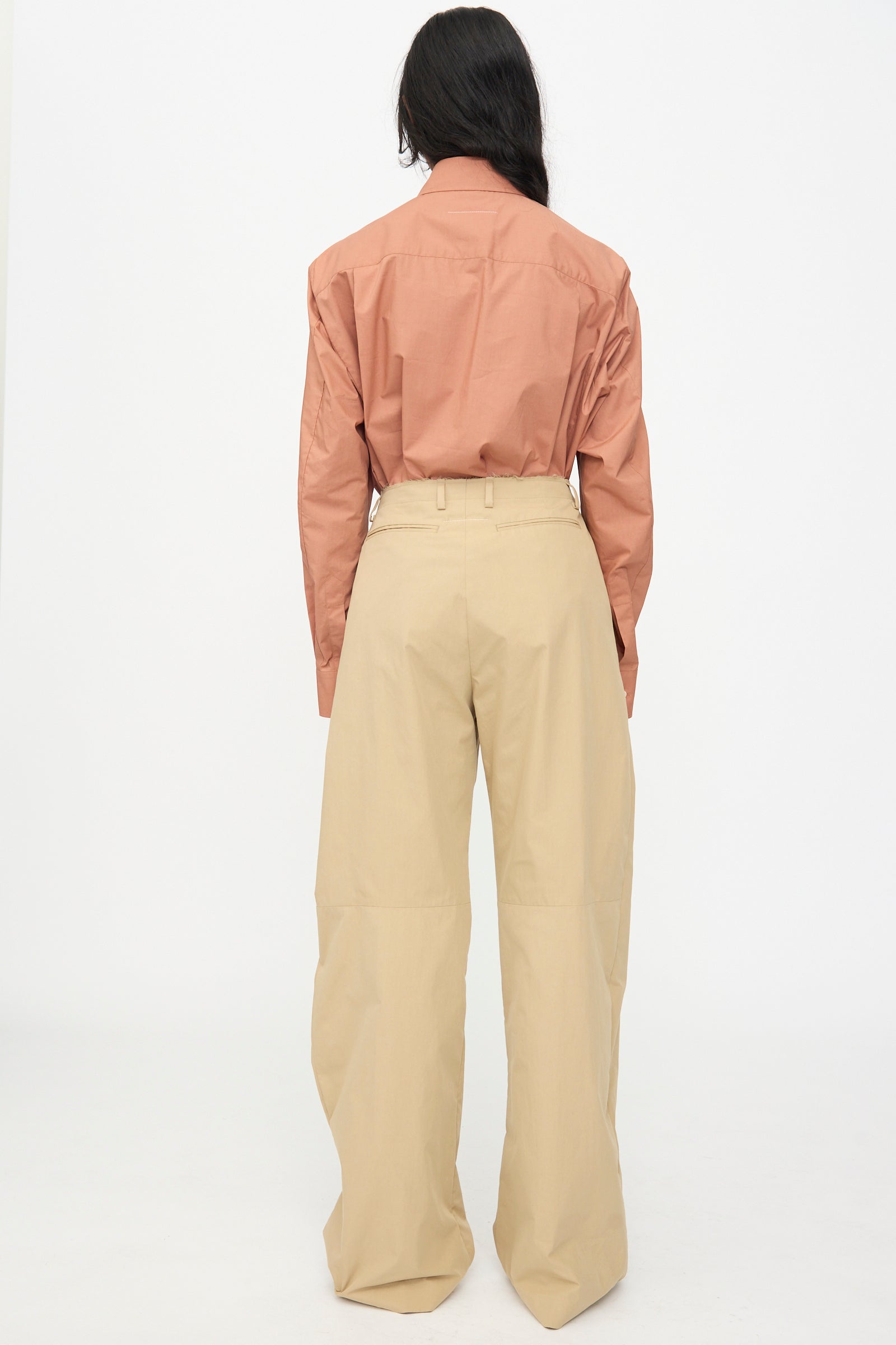 A person stands with their back to the camera, wearing a long-sleeve rust shirt and MM6 Pants in Beige, high-waisted with a relaxed fit wide leg, set against a plain white background.