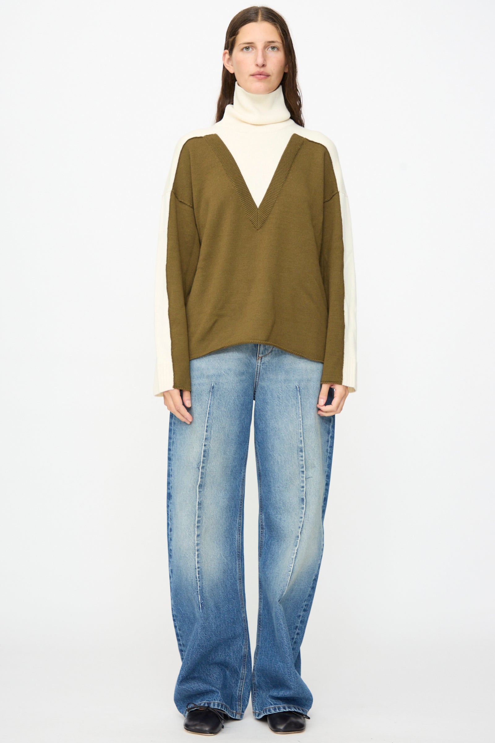 A person stands against a plain background, wearing a two-tone olive and cream sweater with a deep V design, paired with MM6 Pants in Blue—relaxed fit, wide-leg jeans in faded denim—and black shoes.