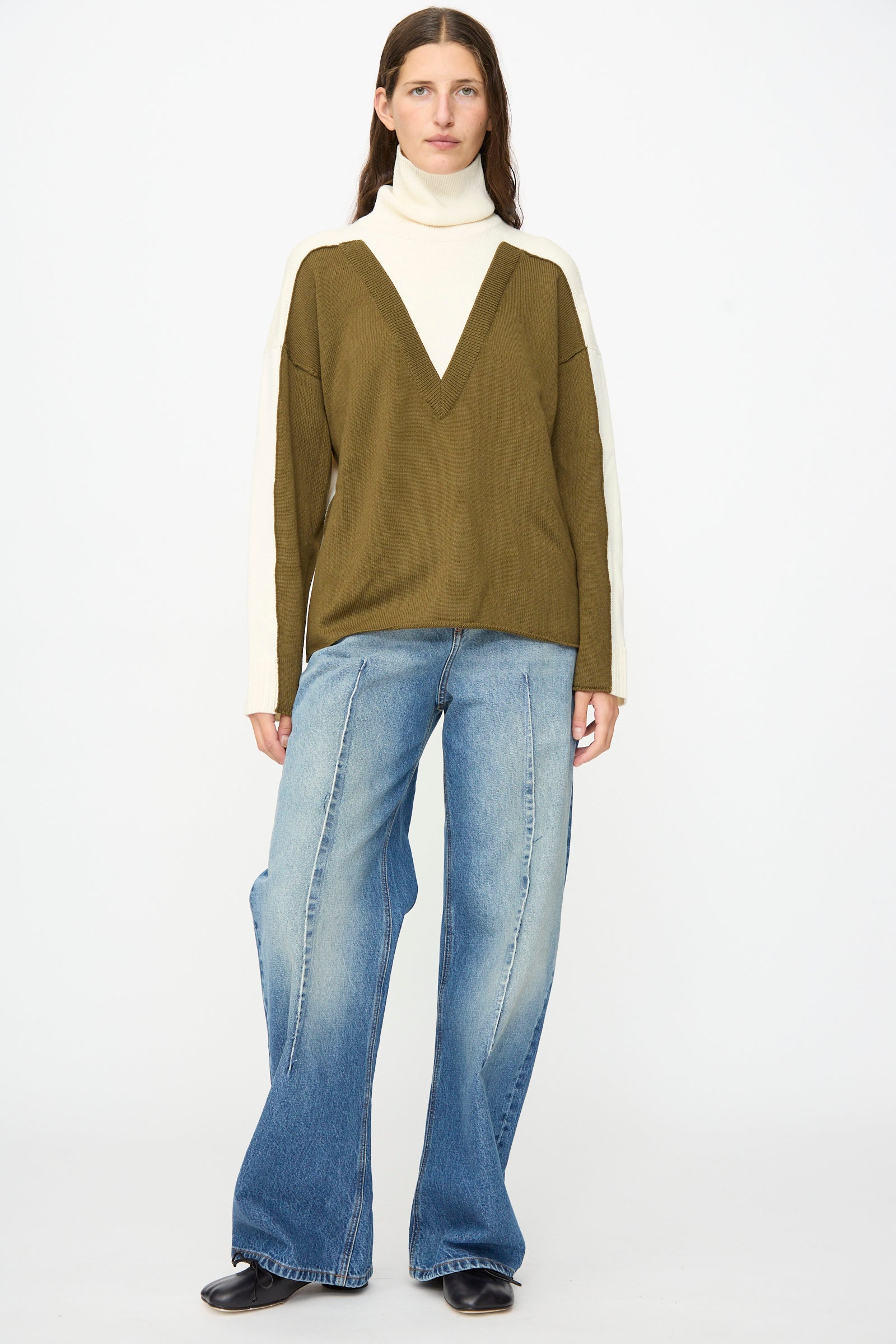 Person wearing a green and cream V-neck sweater over a white turtleneck, paired with MM6 Pants in Blue—relaxed fit, mid-rise denim—along with black shoes, standing against a plain white background.
