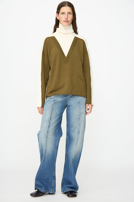 Person wearing a green and cream V-neck sweater over a white turtleneck, paired with MM6 Pants in Blue—relaxed fit, mid-rise denim—along with black shoes, standing against a plain white background.