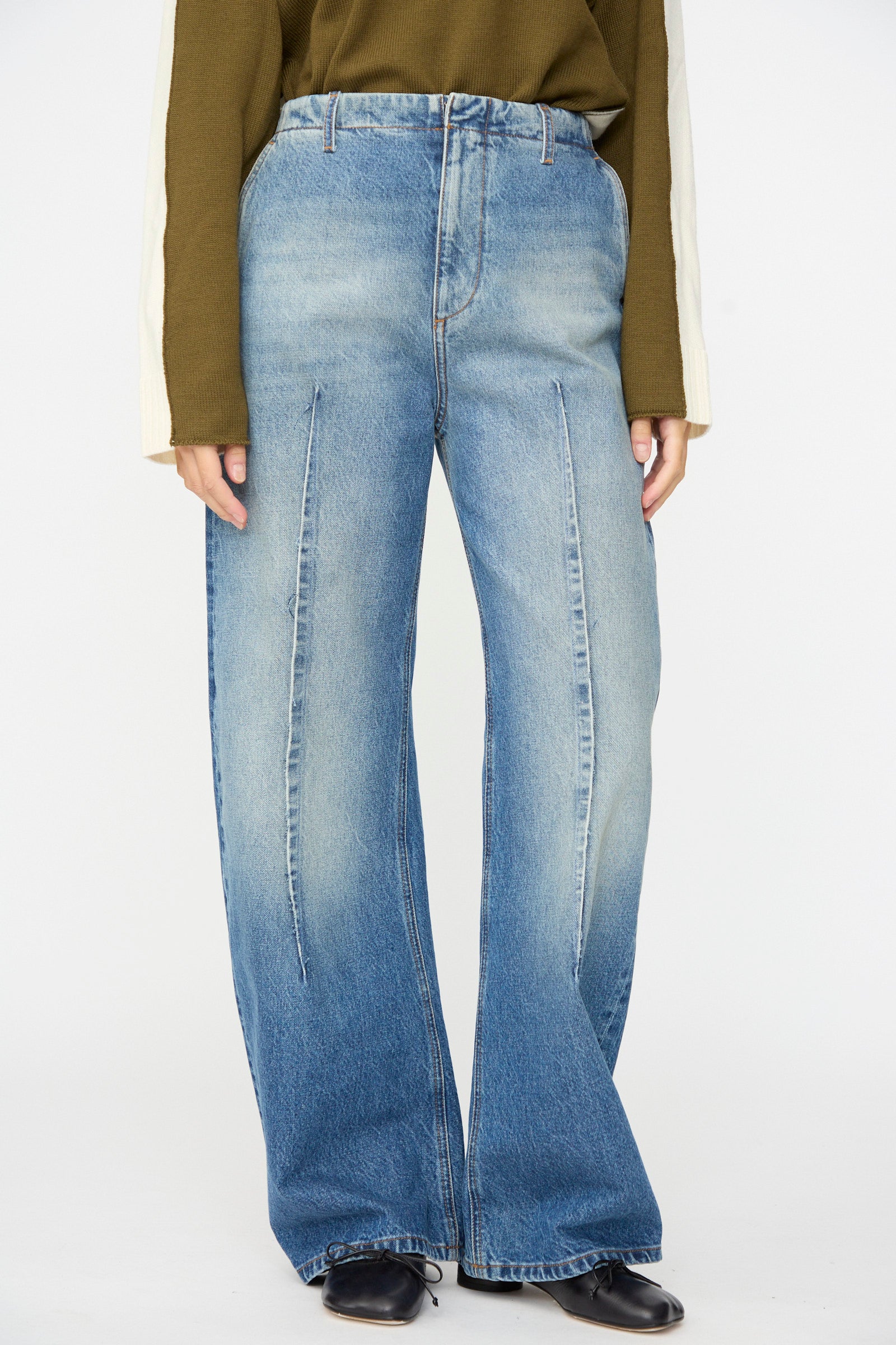 A person wearing MM6 Pants in Blue—relaxed fit, high-waisted, wide-leg jeans with front seam detailing crafted from faded denim—paired with a green long-sleeve top and black shoes, standing against a plain white background.
