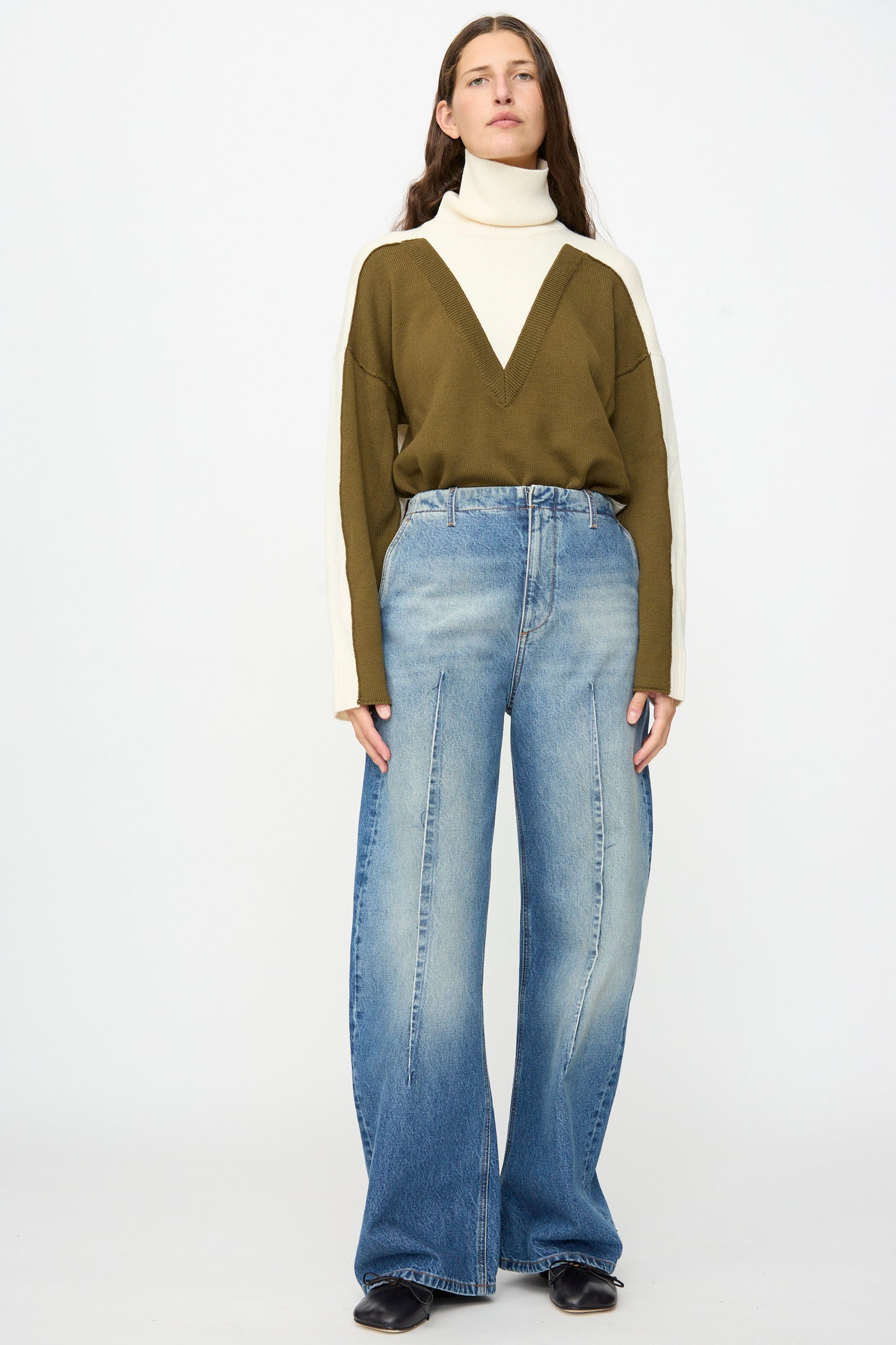 A person stands against a plain background, wearing MM6 Pants in Blue—relaxed fit, wide-leg jeans with visible seams—styled with a white turtleneck, an olive V-pattern sweater, and black shoes.