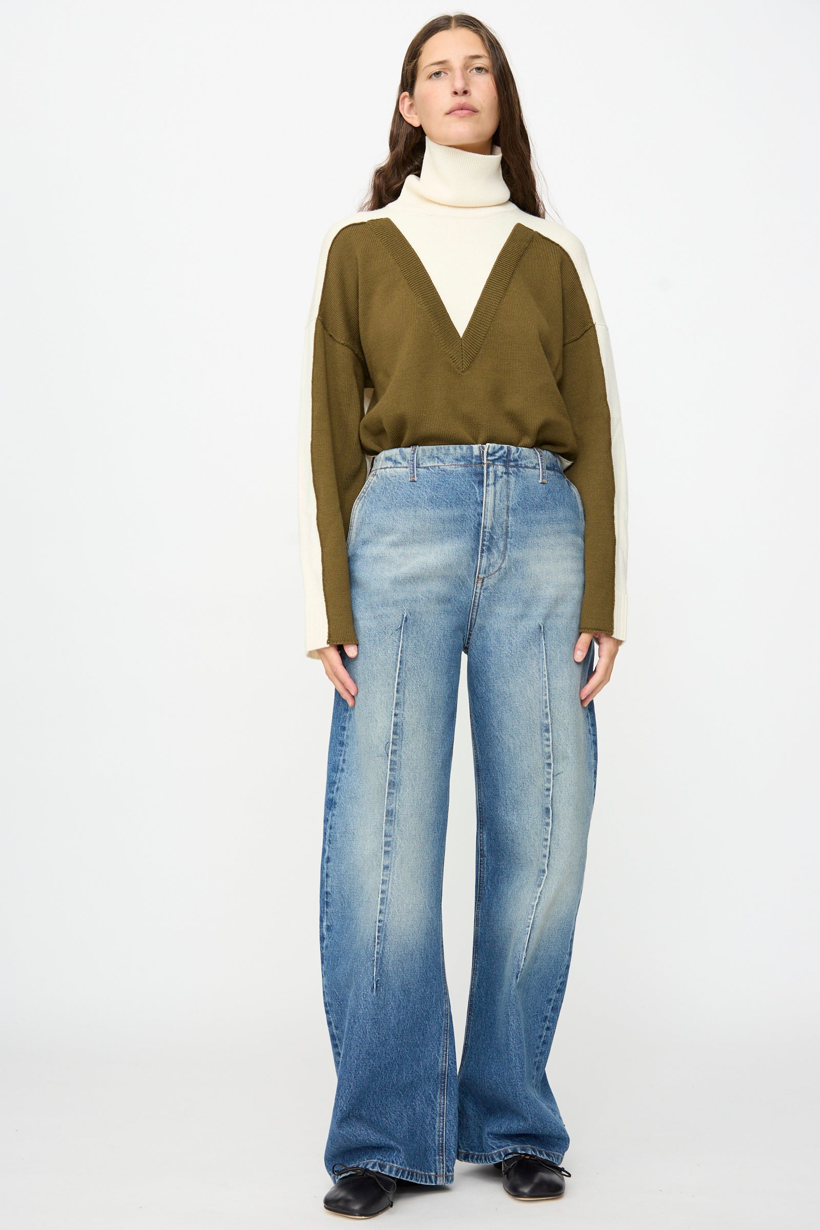 A person stands against a plain background, wearing MM6 Pants in Blue—relaxed fit, wide-leg jeans with visible seams—styled with a white turtleneck, an olive V-pattern sweater, and black shoes.