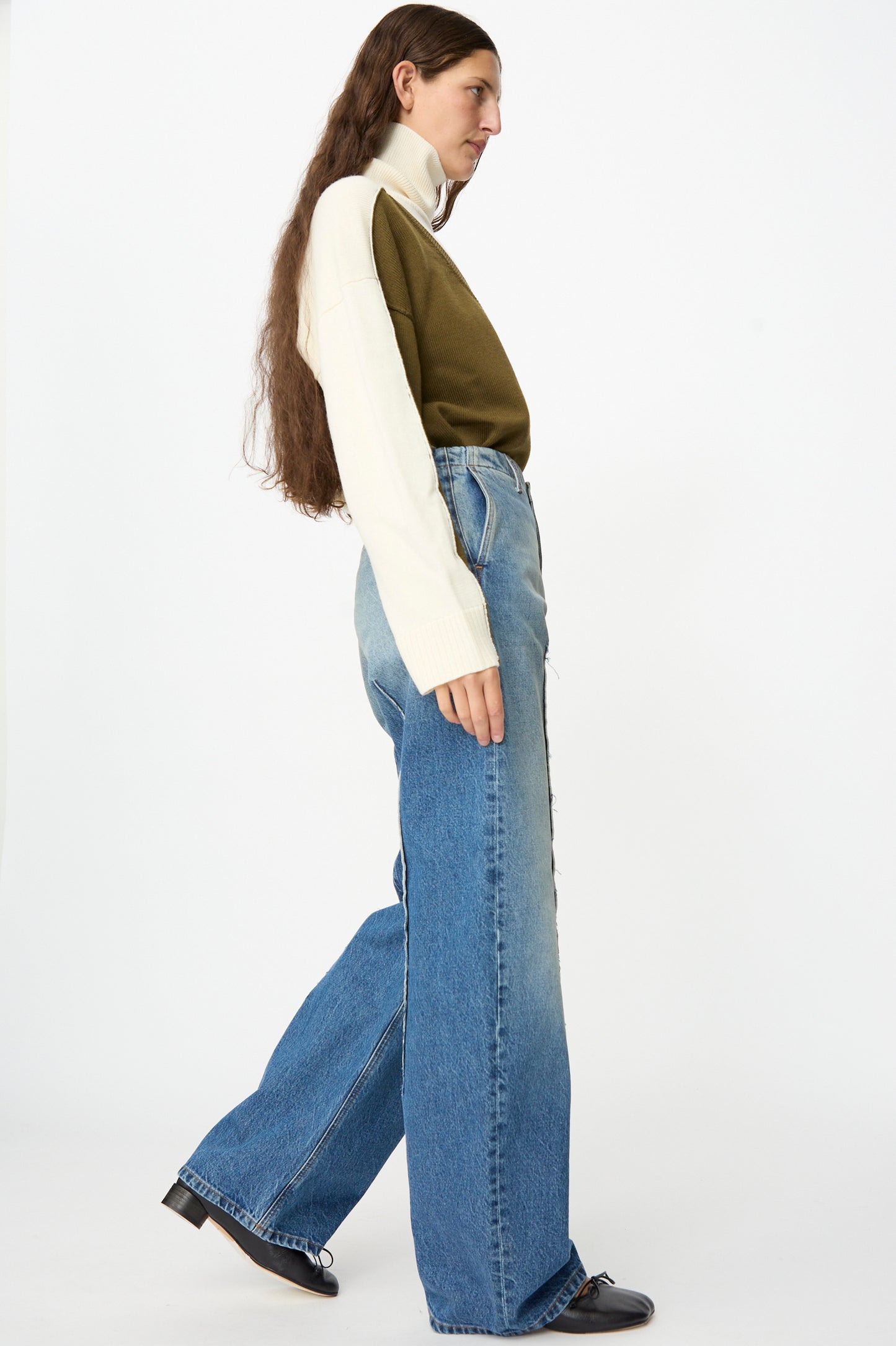 A person with long hair, dressed in a white and olive green sweater, MM6 Pants in Blue (relaxed fit wide-leg jeans in faded denim), and black shoes stands in side profile against a plain white background.
