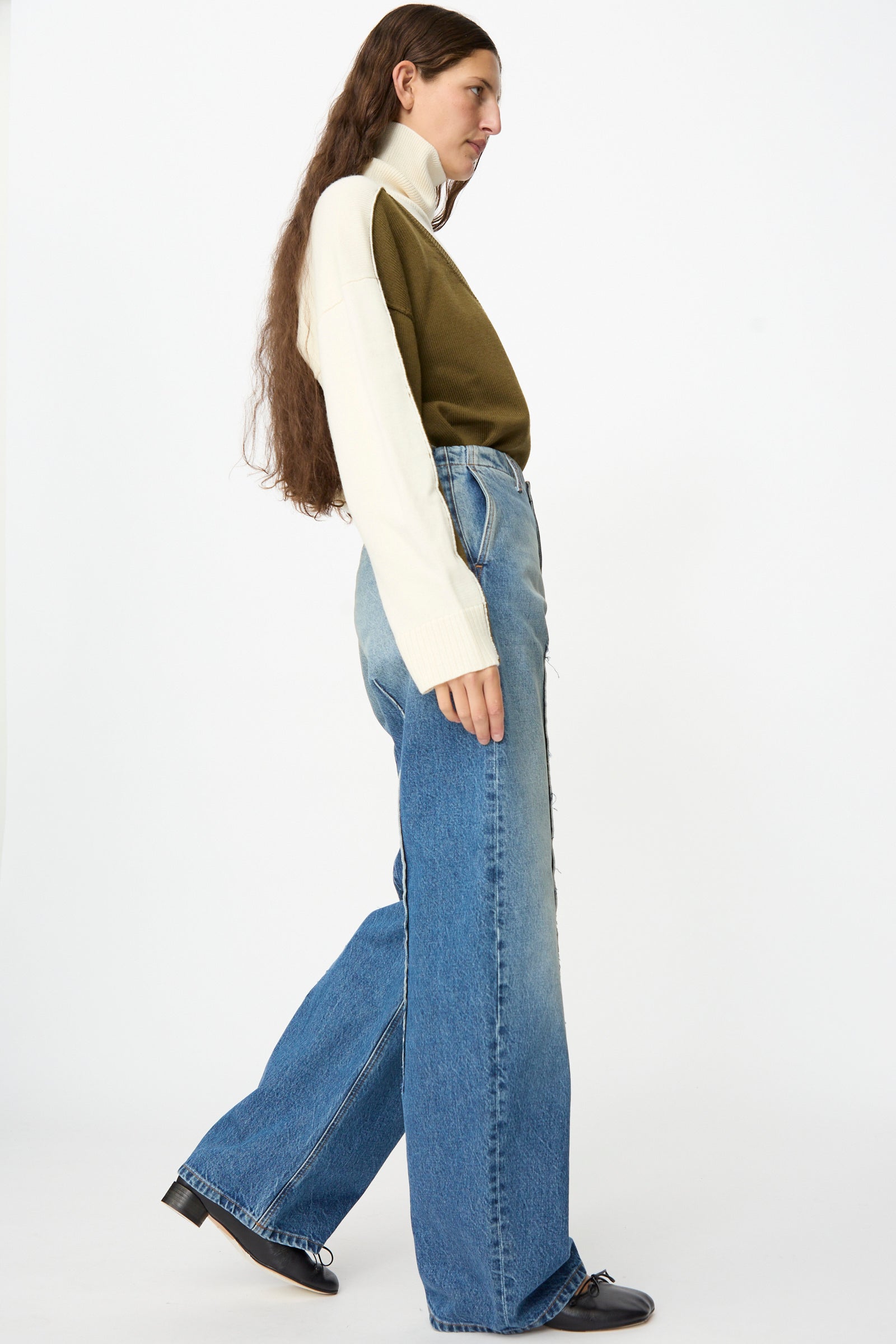 A person with long hair, dressed in a white and olive green sweater, MM6 Pants in Blue (relaxed fit wide-leg jeans in faded denim), and black shoes stands in side profile against a plain white background.