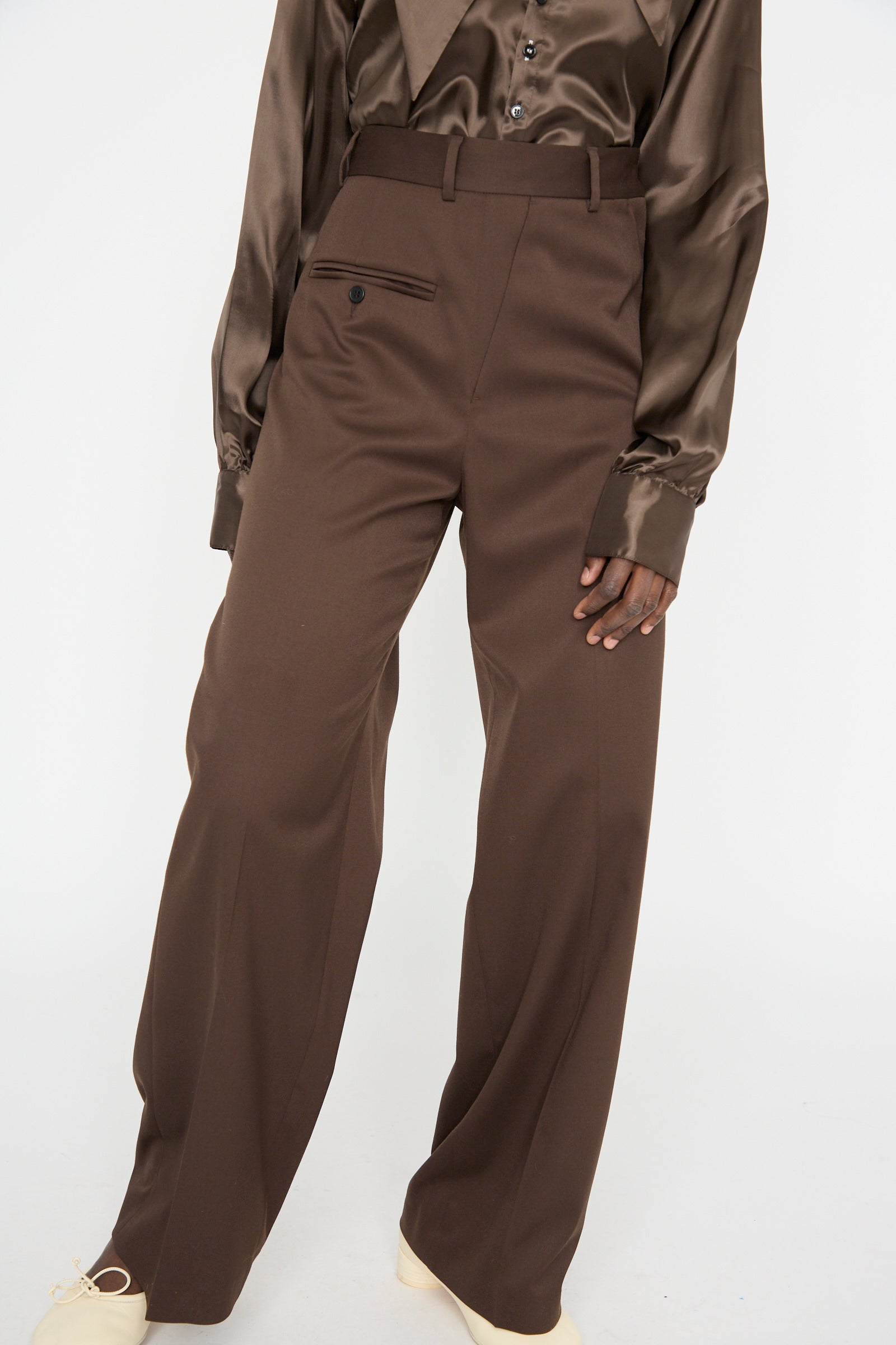 A person stands against a plain background, wearing MM6 Pants in Dark Brown and a relaxed-fit satin blouse.