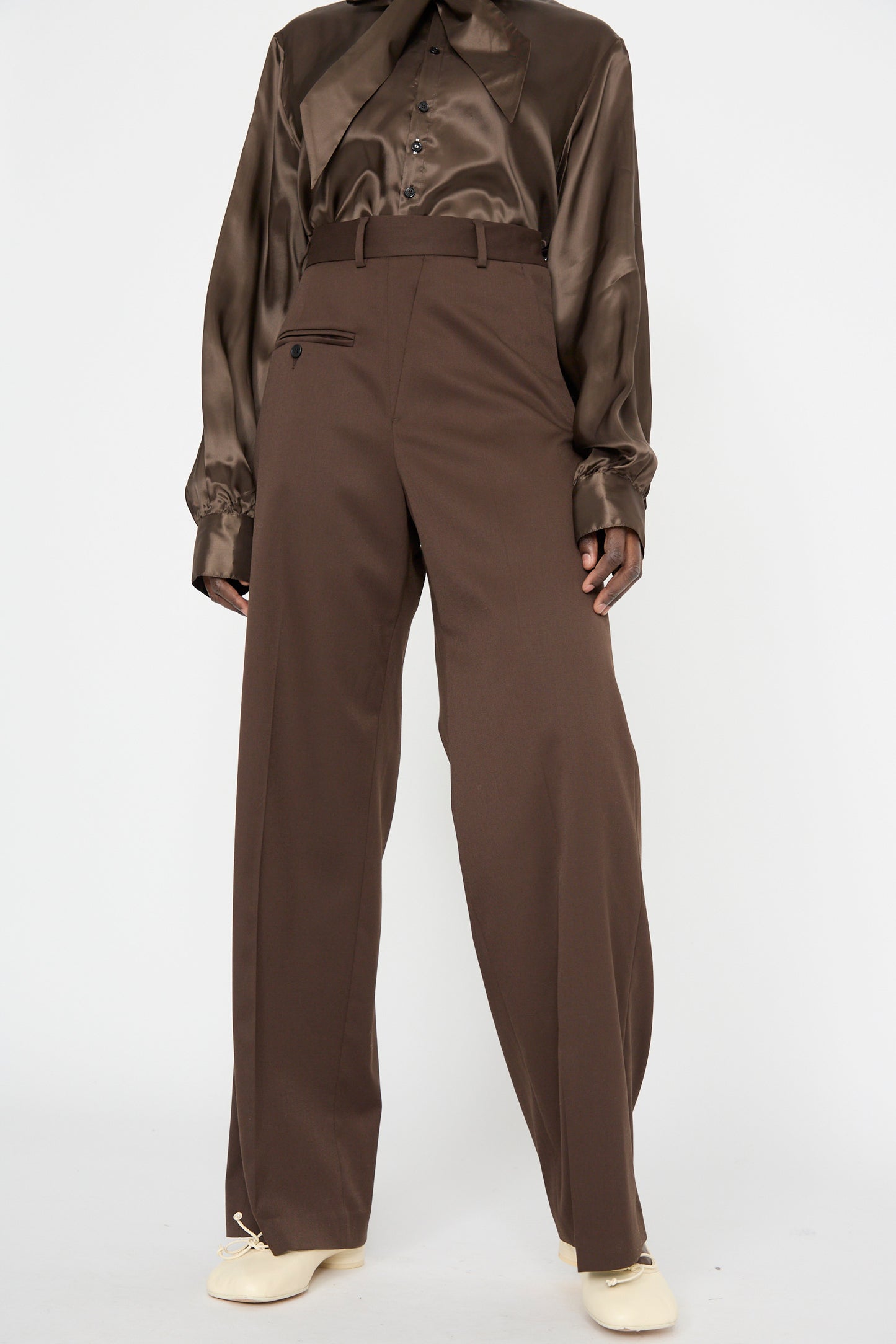 Someone wearing an MM6 Pants in Dark Brown with a glossy brown shirt features a relaxed fit that complements the high-waisted tailored design, complete with button pocket. Cream shoes add subtle elegance to the look.