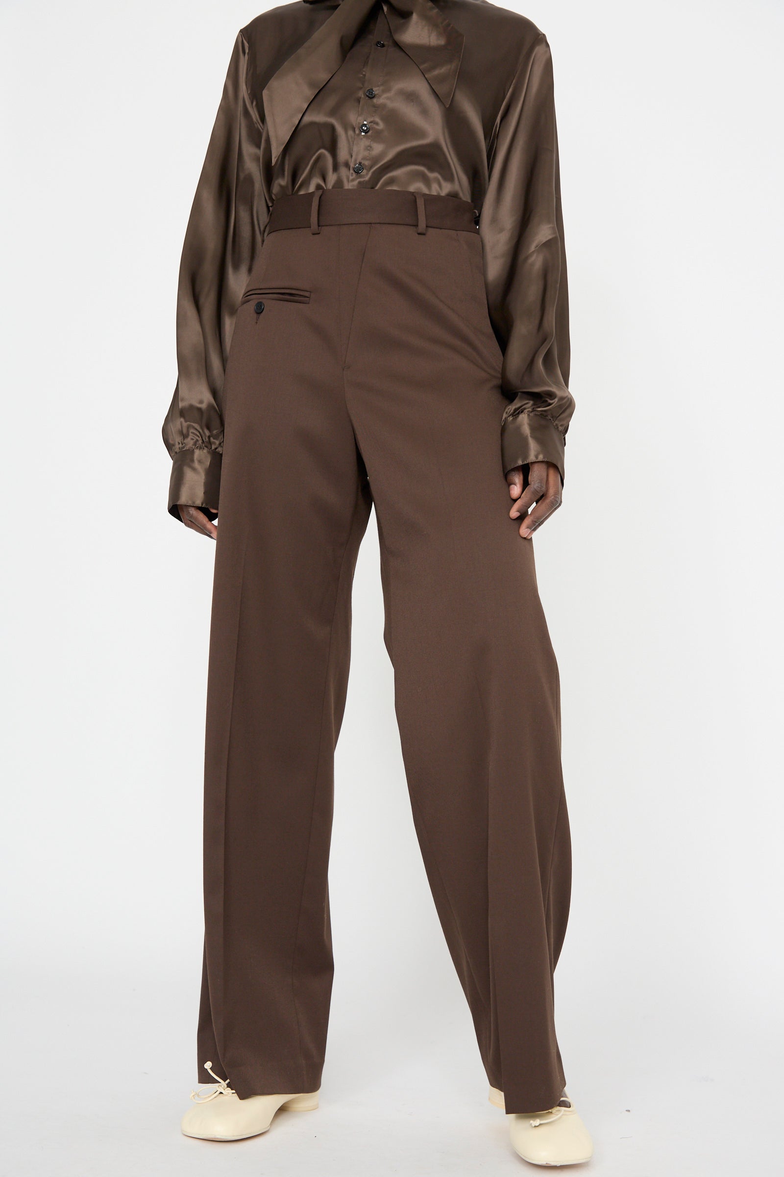 Someone wearing an MM6 Pants in Dark Brown with a glossy brown shirt features a relaxed fit that complements the high-waisted tailored design, complete with button pocket. Cream shoes add subtle elegance to the look.