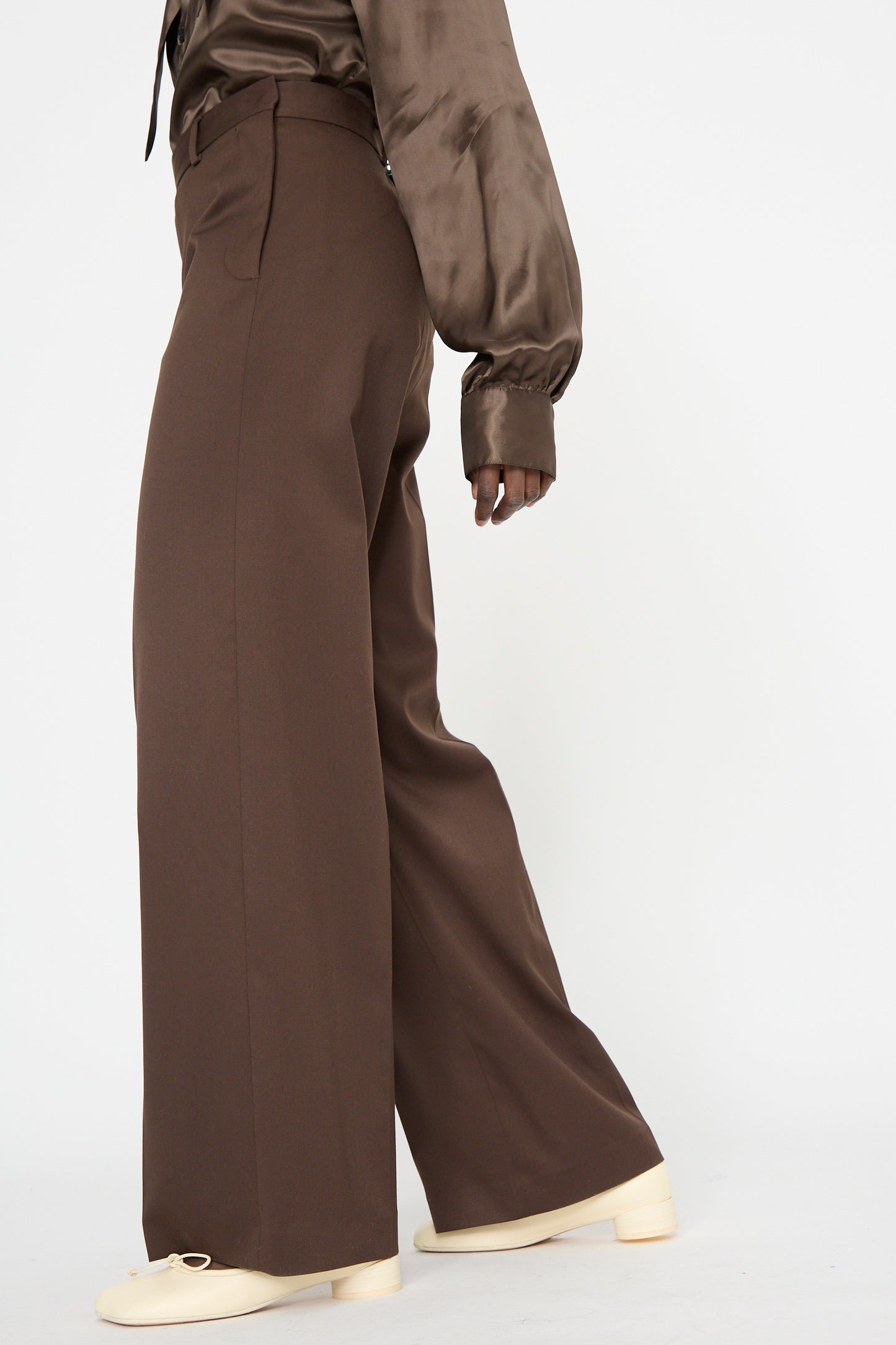 A person in MM6 Pants in Dark Brown and a dark brown satin shirt walks with beige shoes on a plain background, wearing high-waisted wide-leg trousers with a relaxed fit.