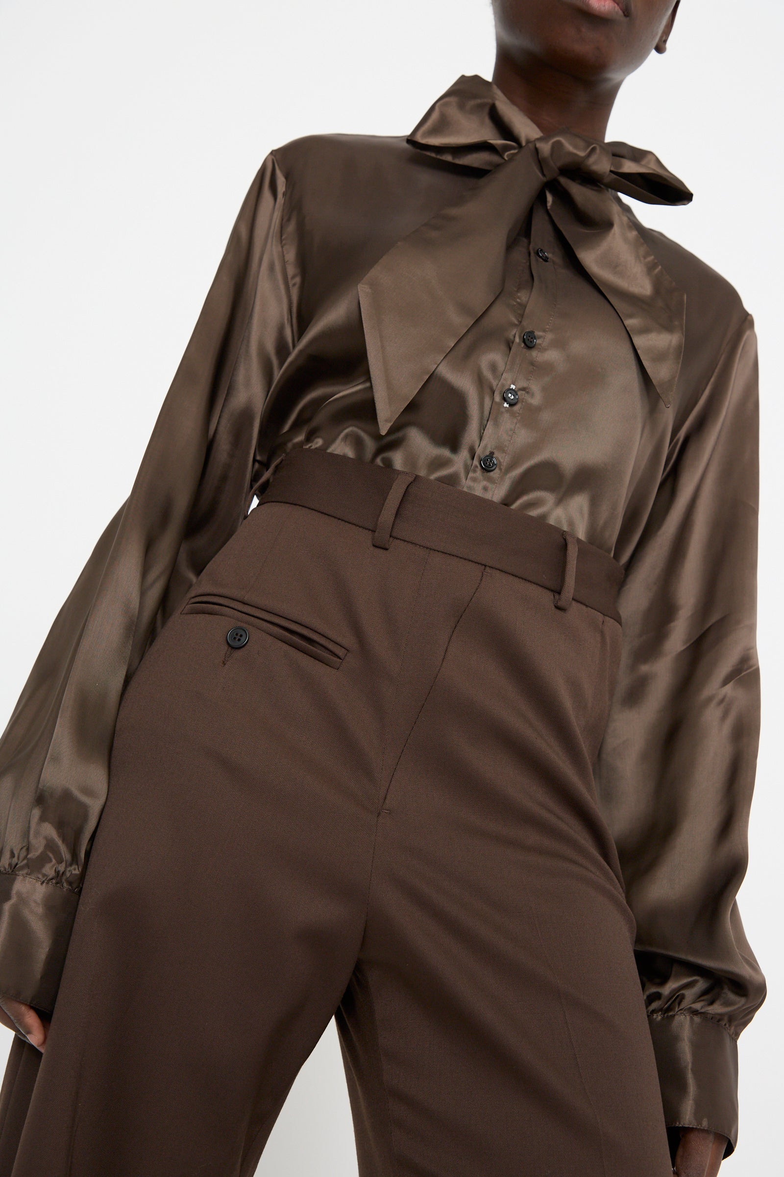 Wearing MM6's silky brown blouse featuring a large bow, the person pairs it elegantly with high-waisted Pants in Dark Brown against a plain background for a stunning look.