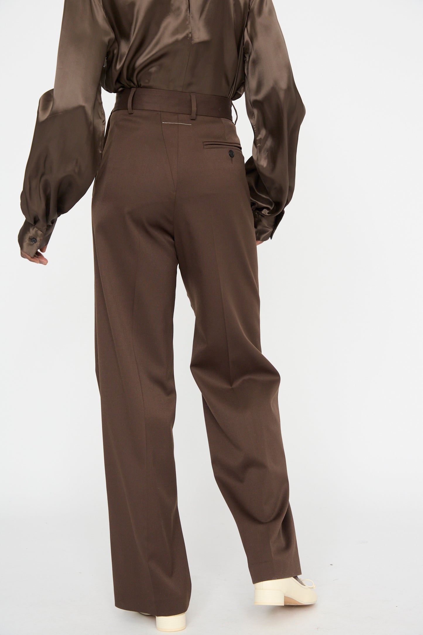 A person is shown from behind wearing a silky blouse and MM6's Dark Brown Pants, which are tailored and high-waisted with a relaxed fit, standing against a plain background.