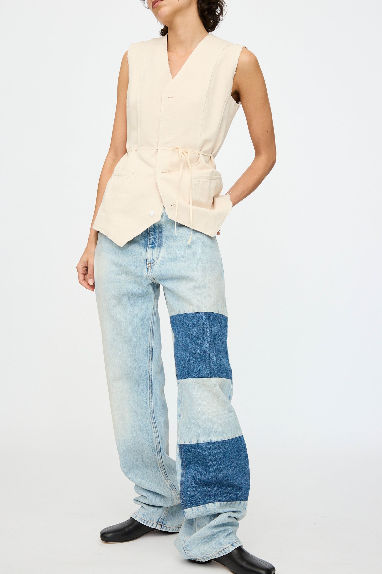 A person wearing a sleeveless beige top with drawstrings and MM6's Pants 5 Pockets, which are loose light blue jeans with a patchwork denim design, paired with black shoes.