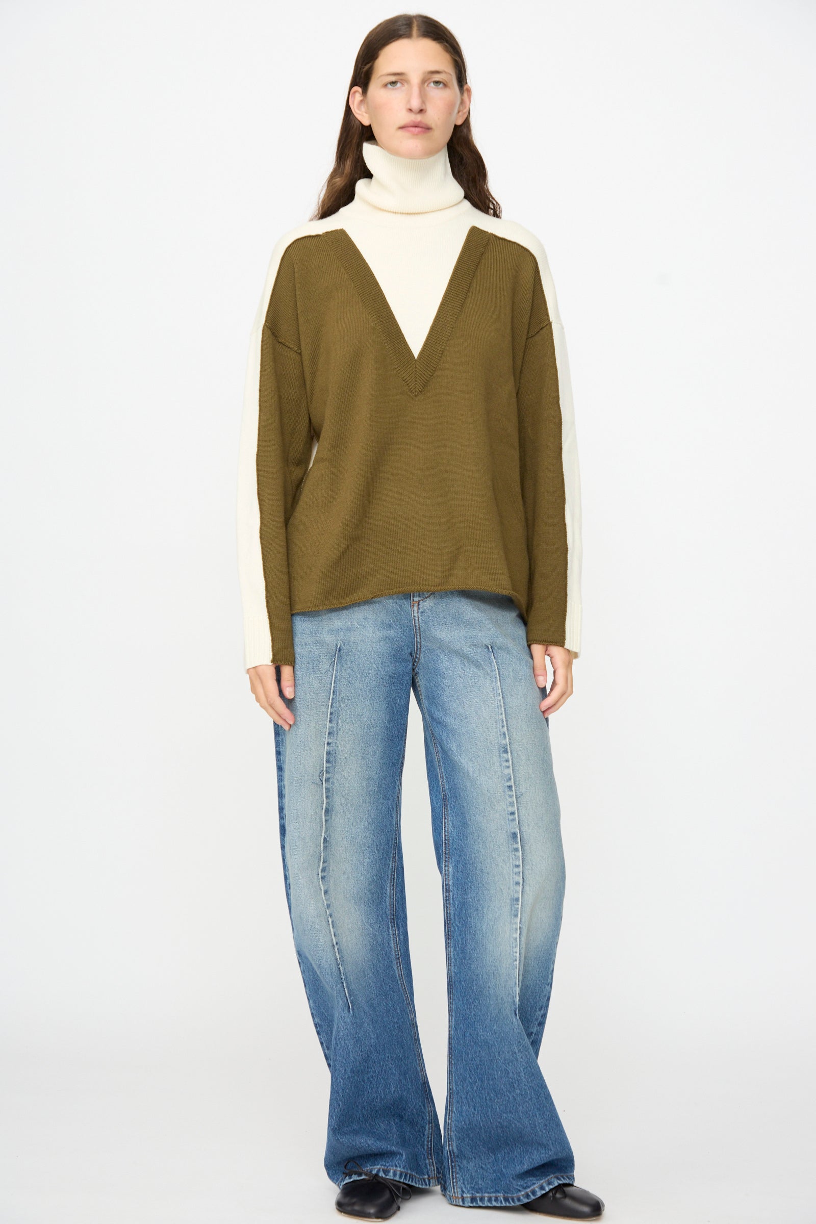A person stands against a white background wearing the MM6 Pullover in Off White-khaki layered over a wool turtleneck, paired with wide-leg blue jeans and black shoes.