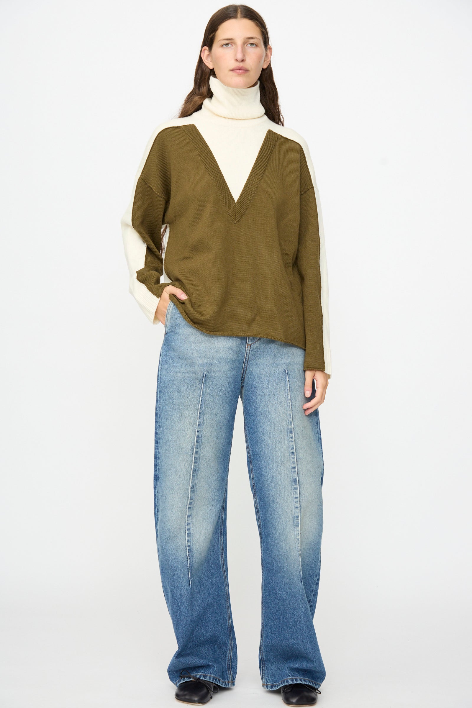 A person wearing the MM6 Pullover in Off White-khaki—a loose, color-blocked wool turtleneck sweater featuring an olive and cream wide V detail—paired with relaxed fit blue jeans and black shoes, standing against a plain white background.