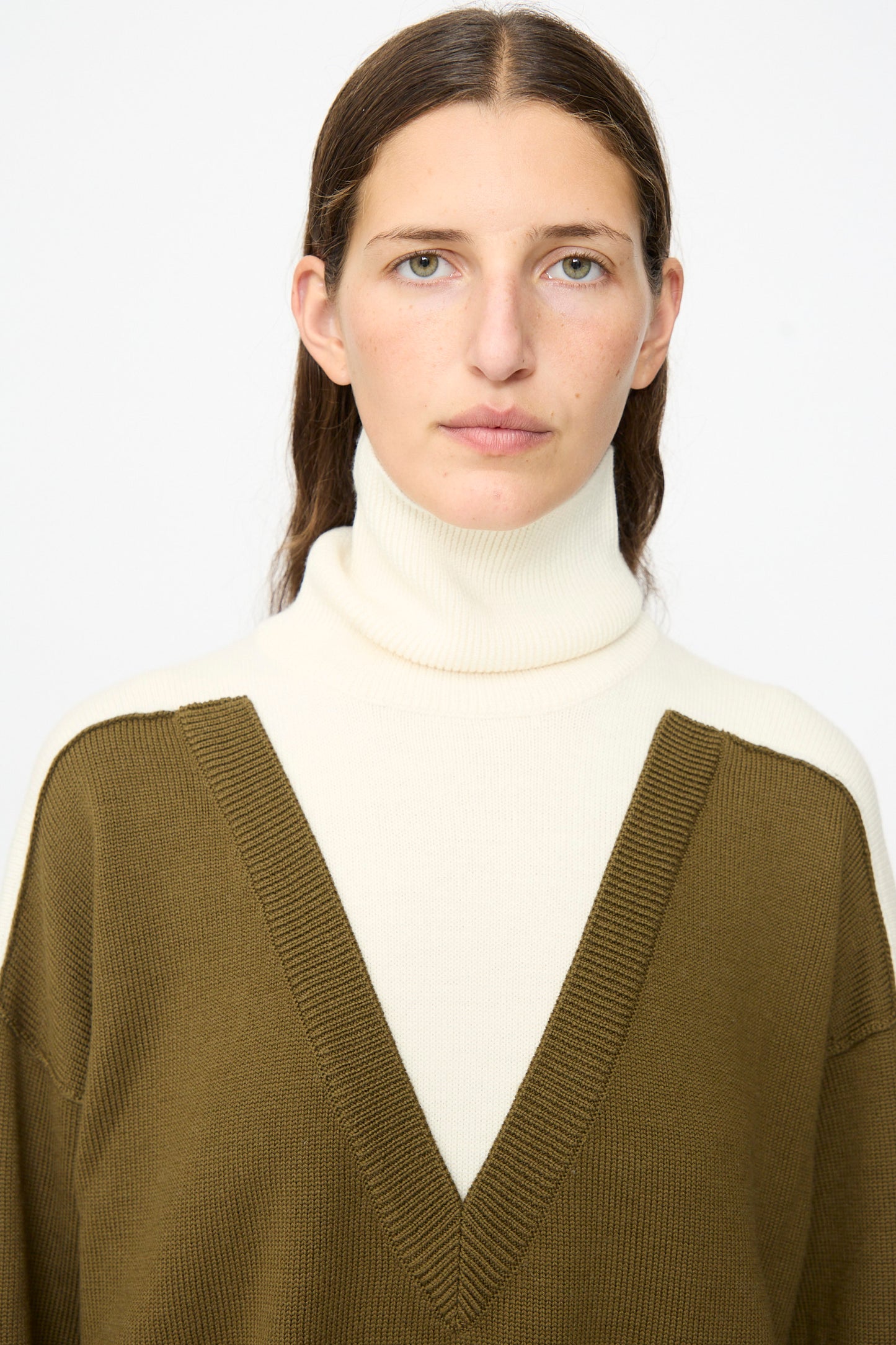 A person with long brown hair wears the MM6 Pullover in Off White-khaki, featuring a white wool turtleneck layered under an olive green V-neck sweater, facing the camera against a plain background.