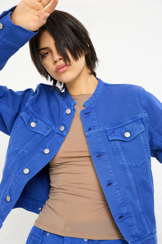 A woman with short hair, wearing a blue MM6 Sports Jacket denim jacket over a tan top, poses with her hand over her forehead.