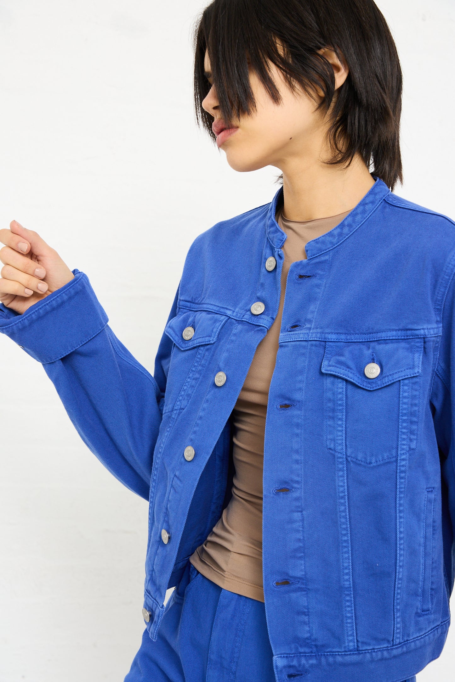 Woman in a blue MM6 Sports Jacket denim over a beige top, looking at her hand, against a white background.