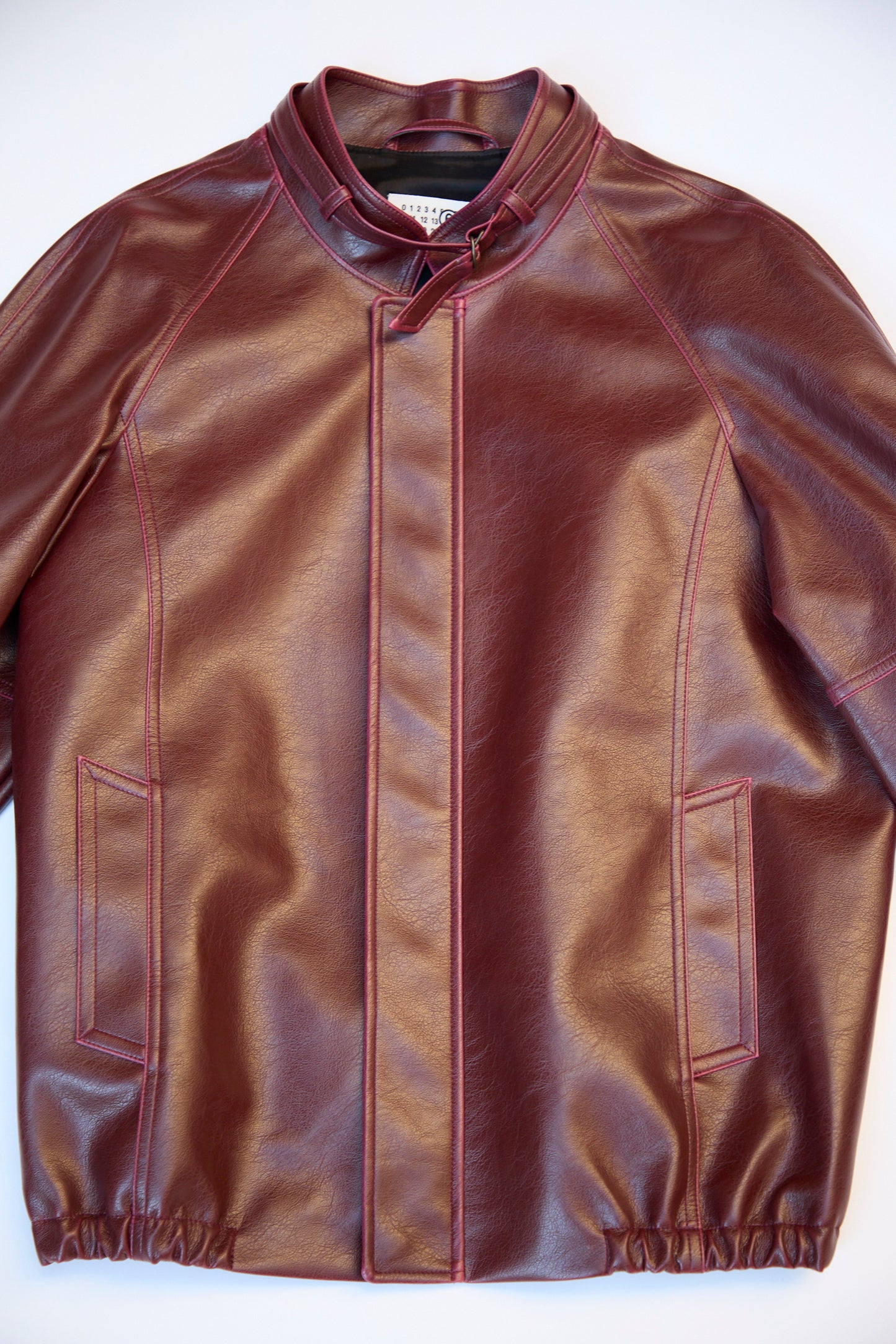 A stylishly displayed MM6 burgundy faux-leather sports jacket, featuring a high collar, front zipper closure, and two front pockets, lies on a flat surface.