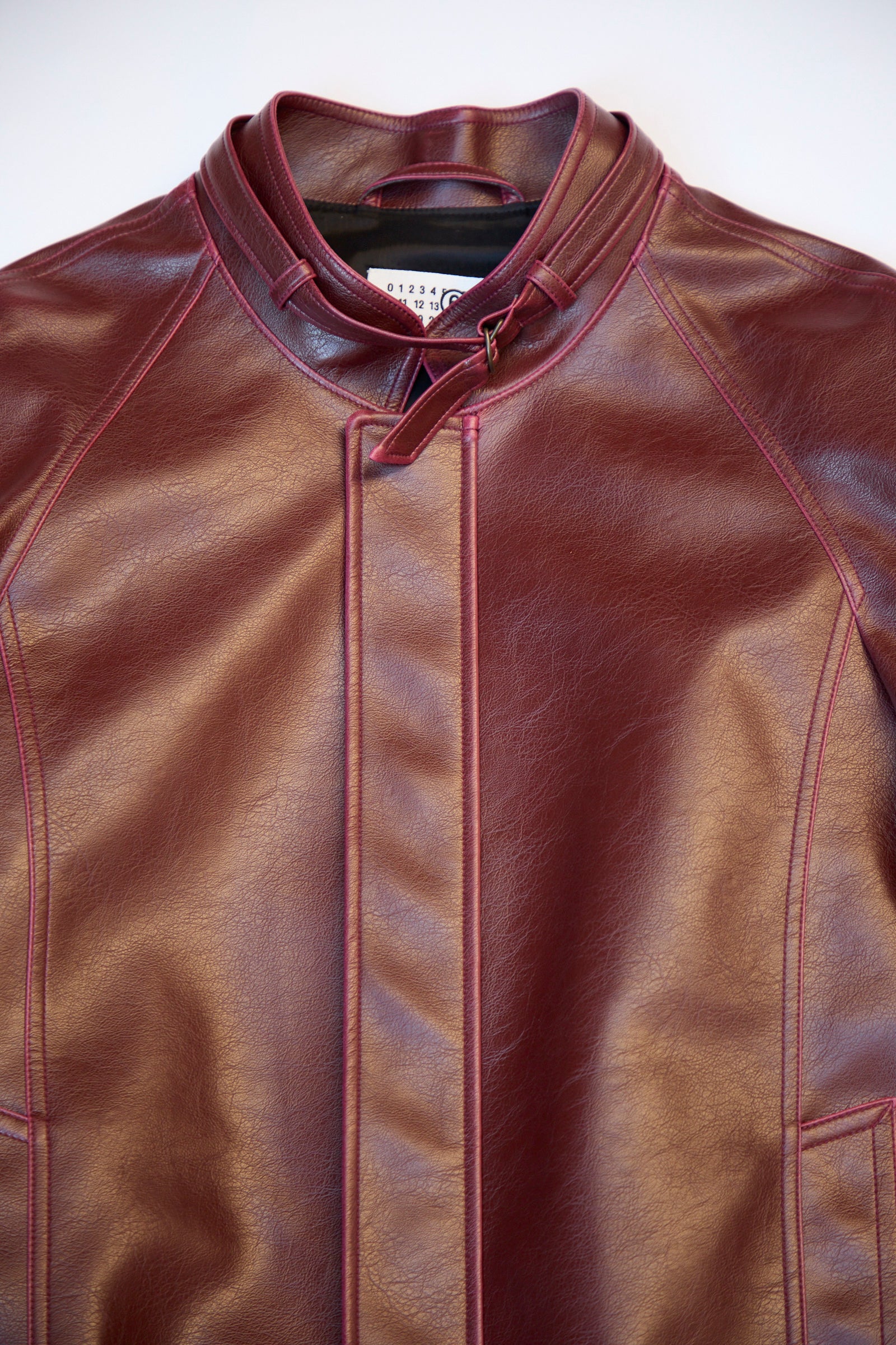 Close-up of an MM6 Sports Jacket in maroon faux-leather, featuring a high collar, front detailing, and visible stitching against a light background.