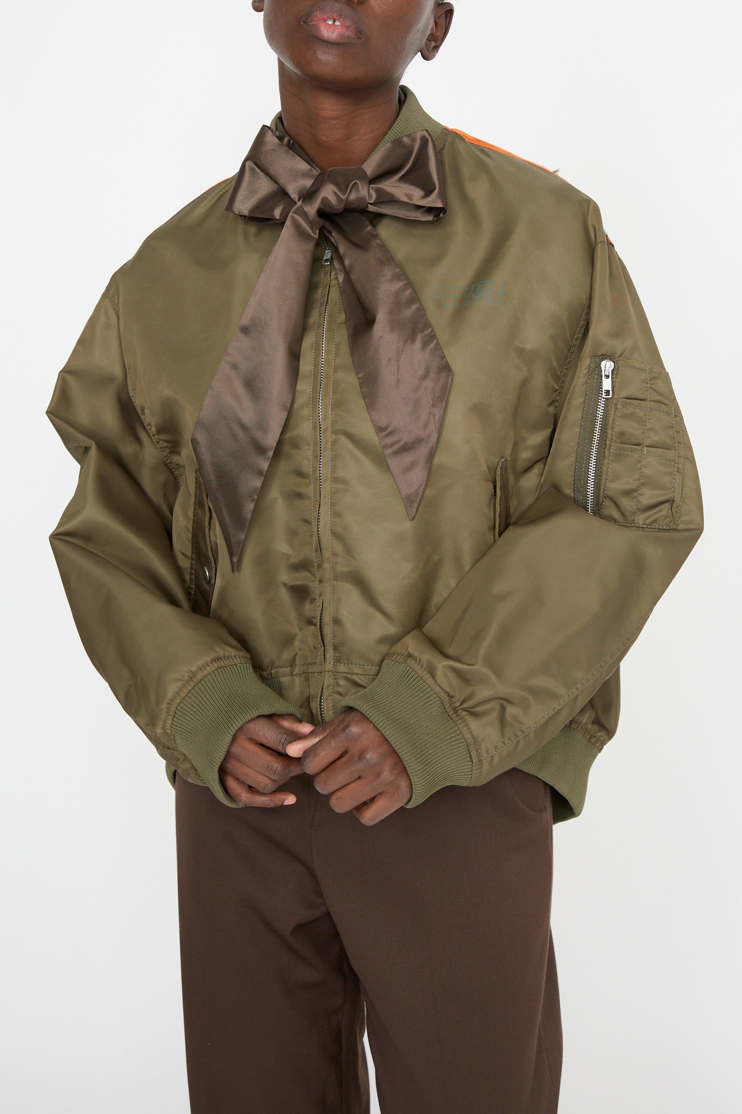 Modeling a Sport Jacket by MM6, an oversized olive green bomber jacket crafted from lightweight polyamide, is styled with brown pants. The look features a large brown bow at the neck and subtle color-block panel design against a plain backdrop.